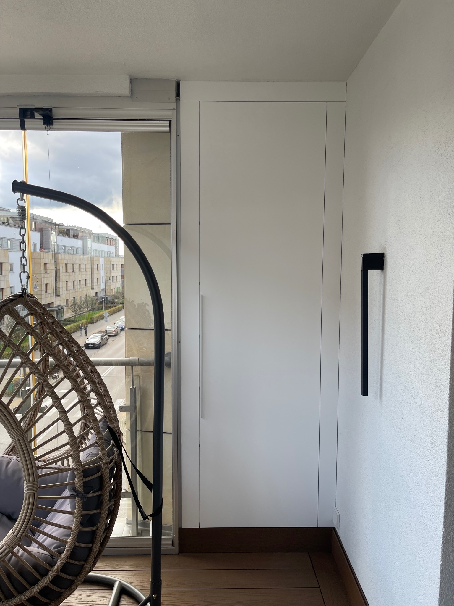 Biała szafa wnękowa z długim uchwytem na balkonie, widok na miasto przez okno, obok wiszące krzesło z poduszkami. Minimalistyczny design.