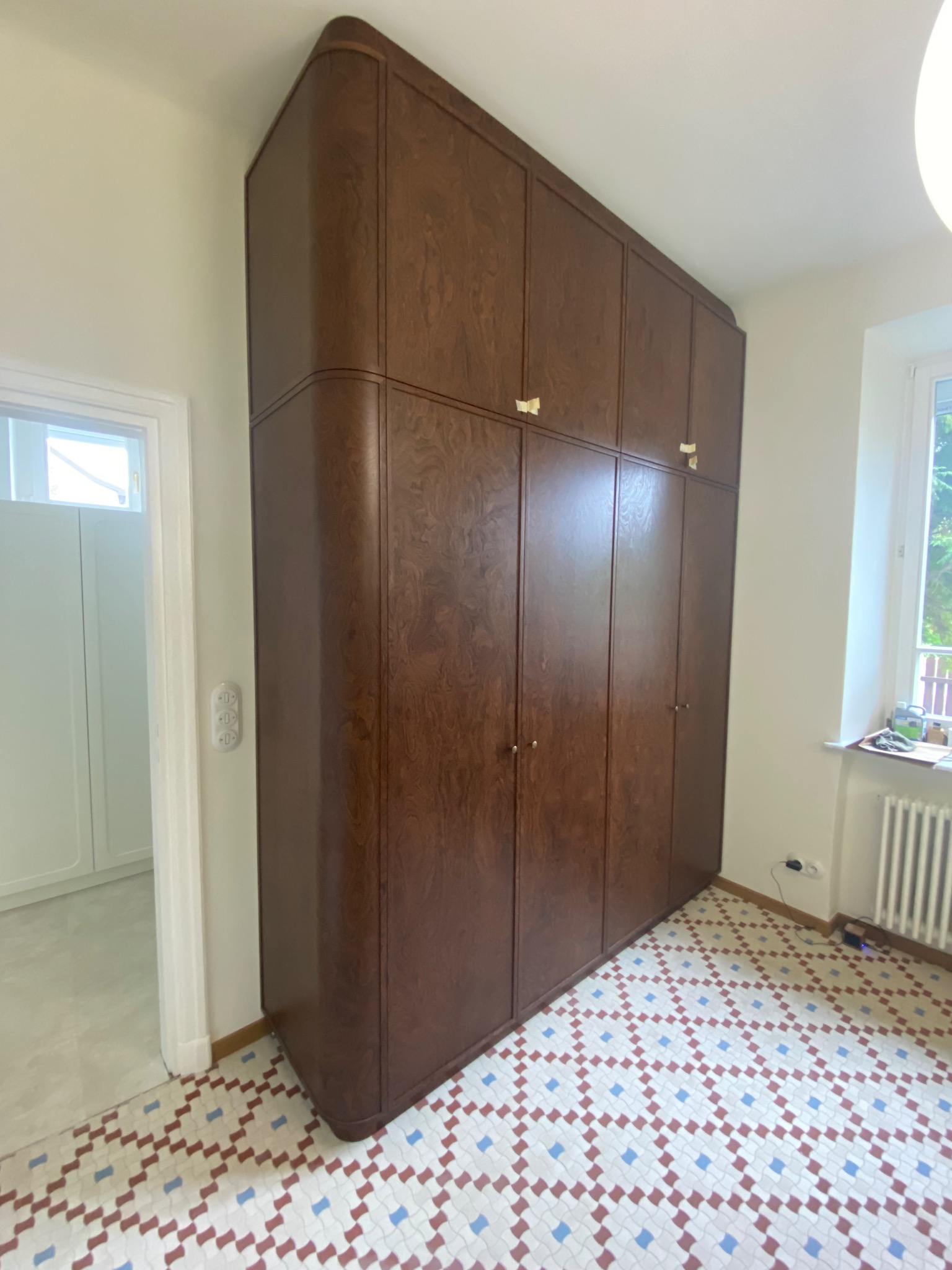 Duża, brązowa szafa z zaokrąglonymi krawędziami, dopasowana do wnętrza z wzorzystą podłogą. Widoczny fragment pokoju z oknem i grzejnikiem.