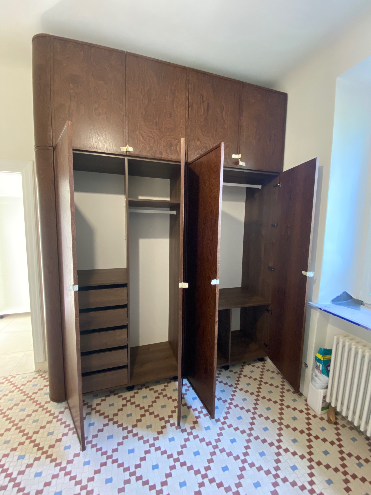 Brązowa szafa wnękowa z otwartymi drzwiami, ukazująca półki i szuflady. Widoczna mozaikowa podłoga i fragment grzejnika. Za drzwiami szafy widoczne wnętrze pomieszczenia.