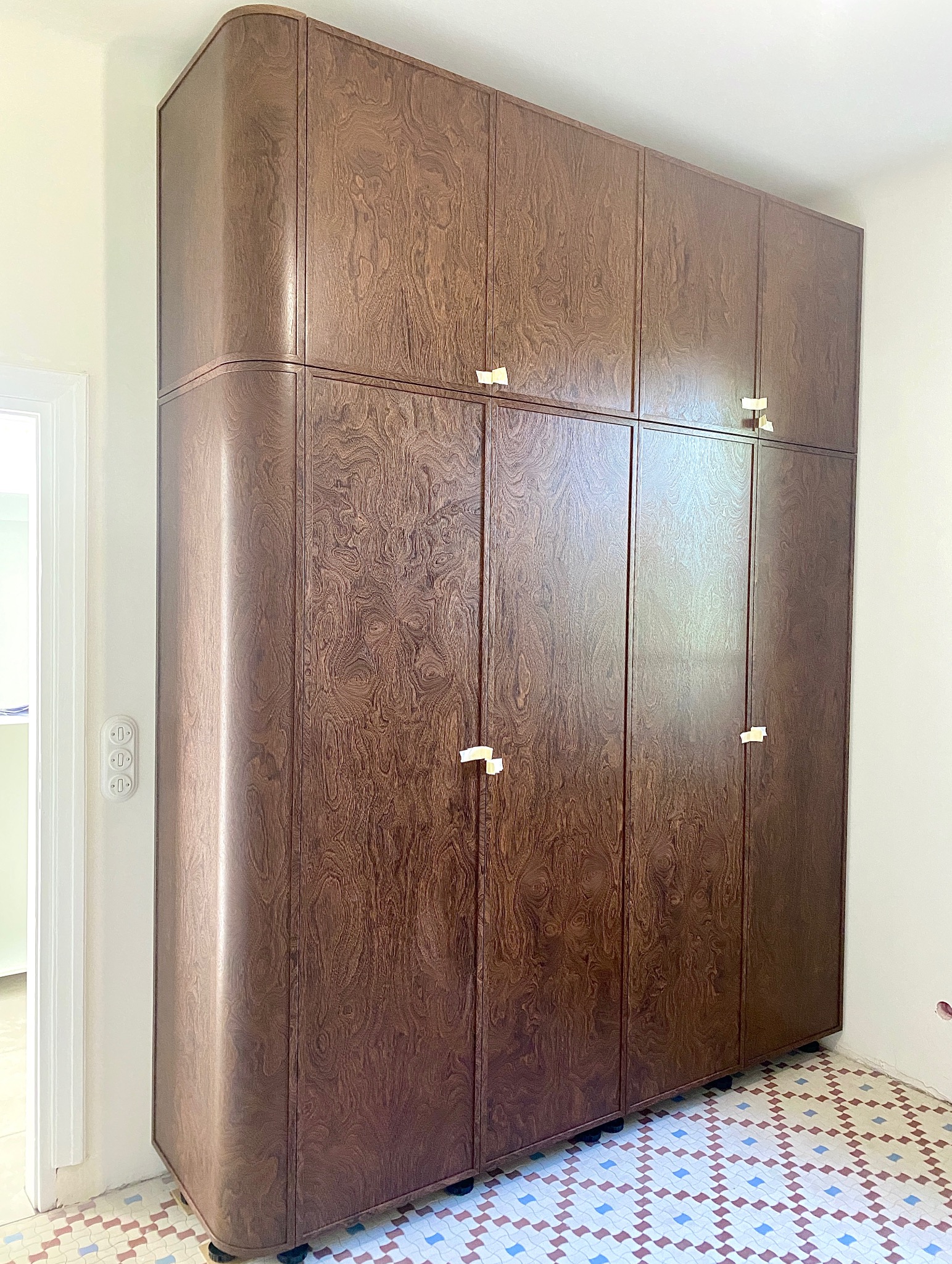 Wysoka, brązowa szafa z zaokrąglonym bokiem i złotymi uchwytami, stojąca na kafelkach w stylu retro. Widoczny fragment ściany z włącznikami.