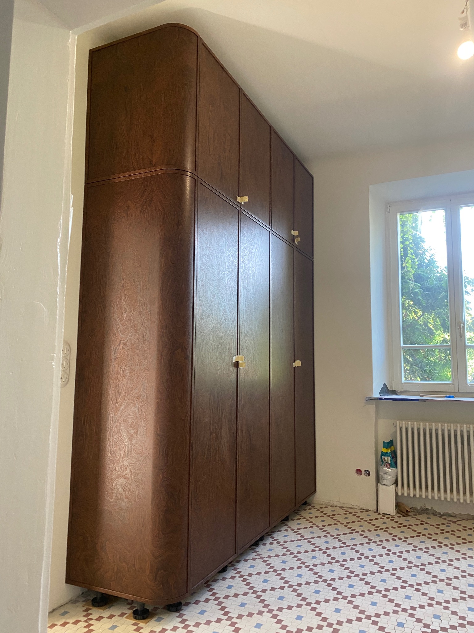 Retro szafa wnękowa z zaokrąglonymi krawędziami i złotymi uchwytami, na tle mozaikowej podłogi i okna z widokiem na zieleń. Wykończenie w odcieniach brązu.