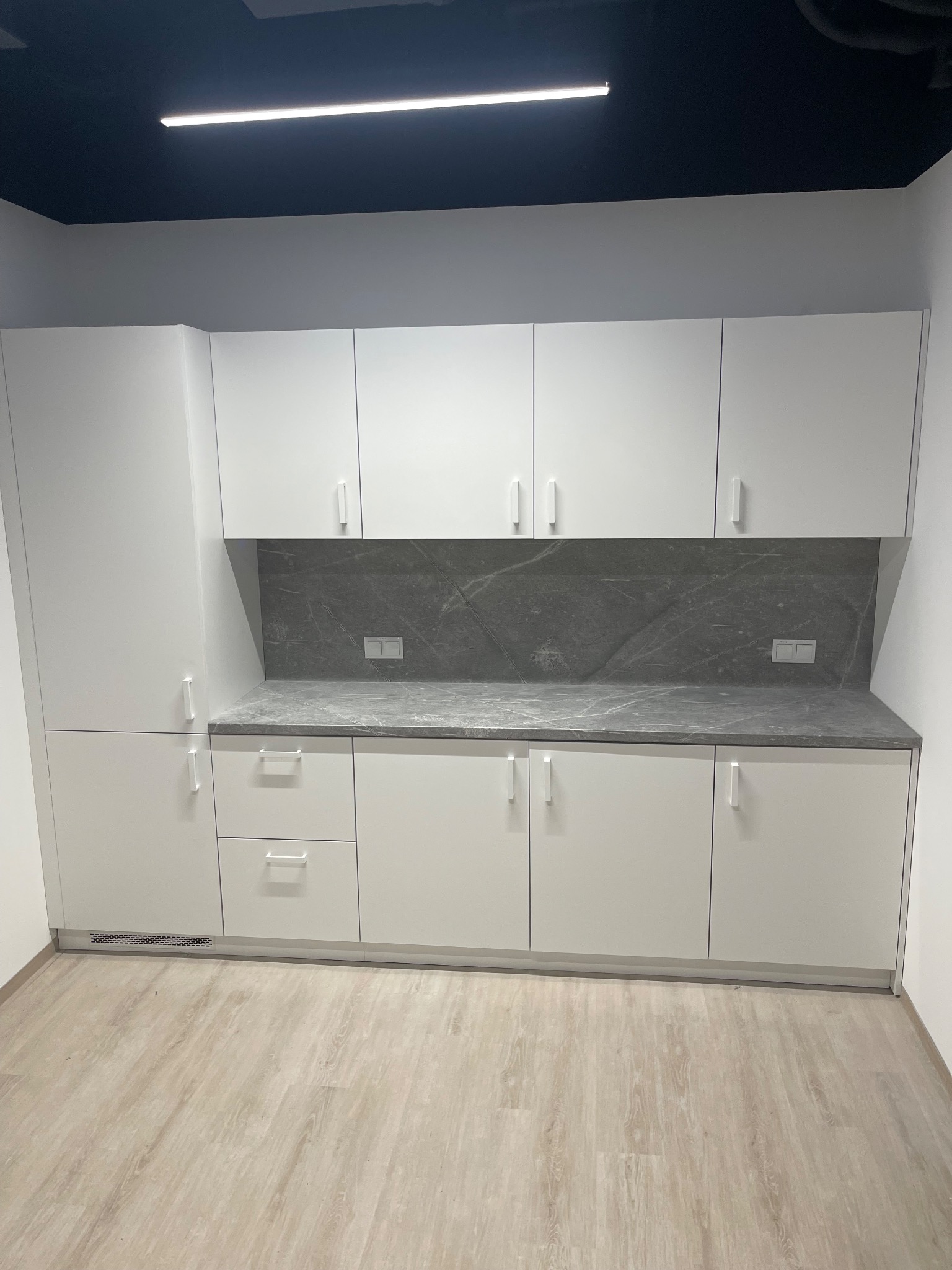 Minimalistyczna, biała zabudowa kuchenna z szarym blatem i marmurowym panelem nad blatem. Widoczne gniazdka elektryczne. Jasna podłoga imitująca drewno.