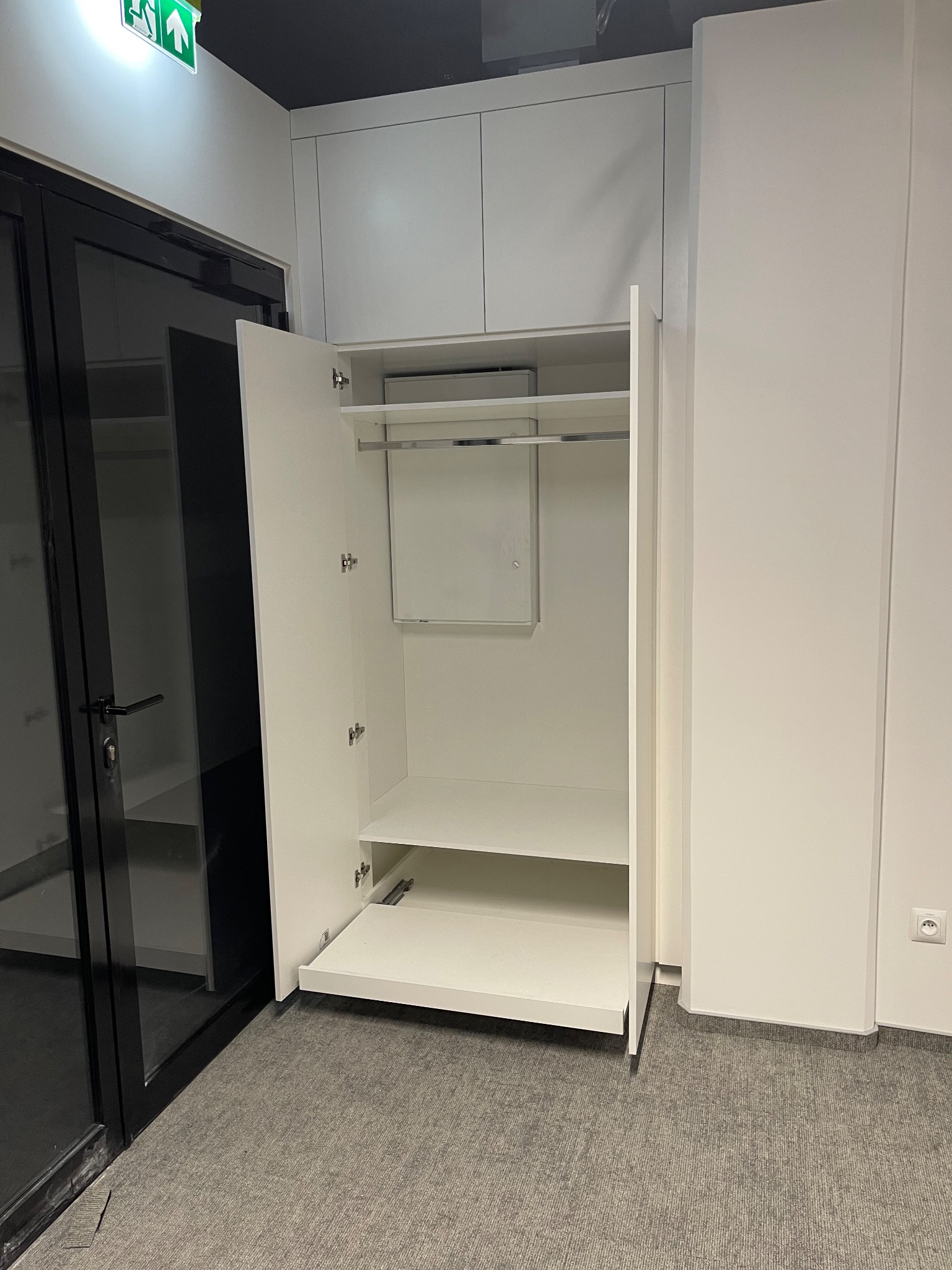 Biała szafa wnękowa z otwartymi drzwiami, widoczne półki i drążek na ubrania. Minimalistyczny design, szara wykładzina podłogowa, czarne obramowanie drzwi.