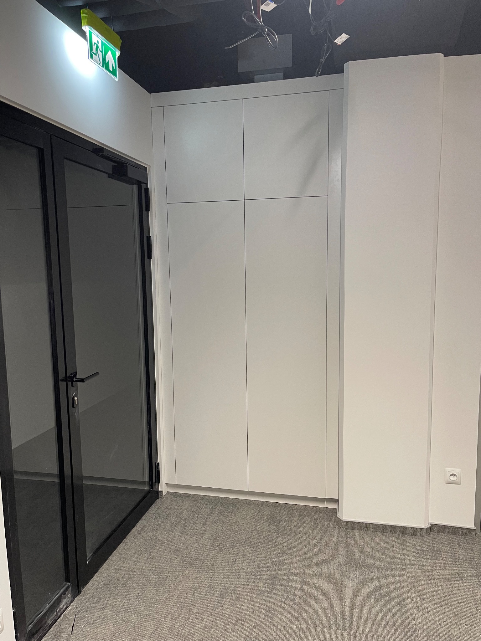 Biała szafa wnękowa z czterema drzwiami, wkomponowana w ścianę, widoczna z perspektywy korytarza z szarym dywanem i czarnymi drzwiami.