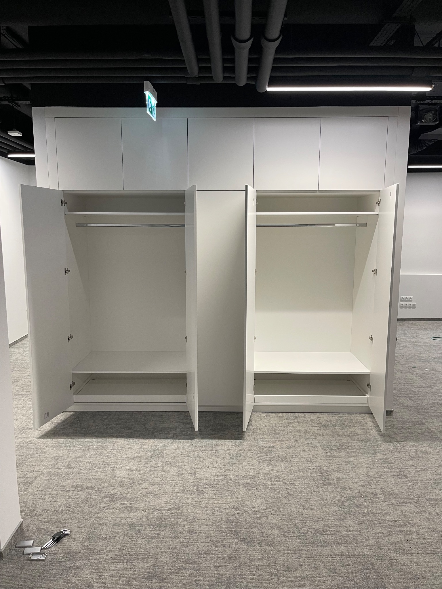 Biała szafa wnękowa z otwartymi drzwiami, widoczne półki i drążek na ubrania. Na podłodze leżą narzędzia. Sufit z instalacjami.