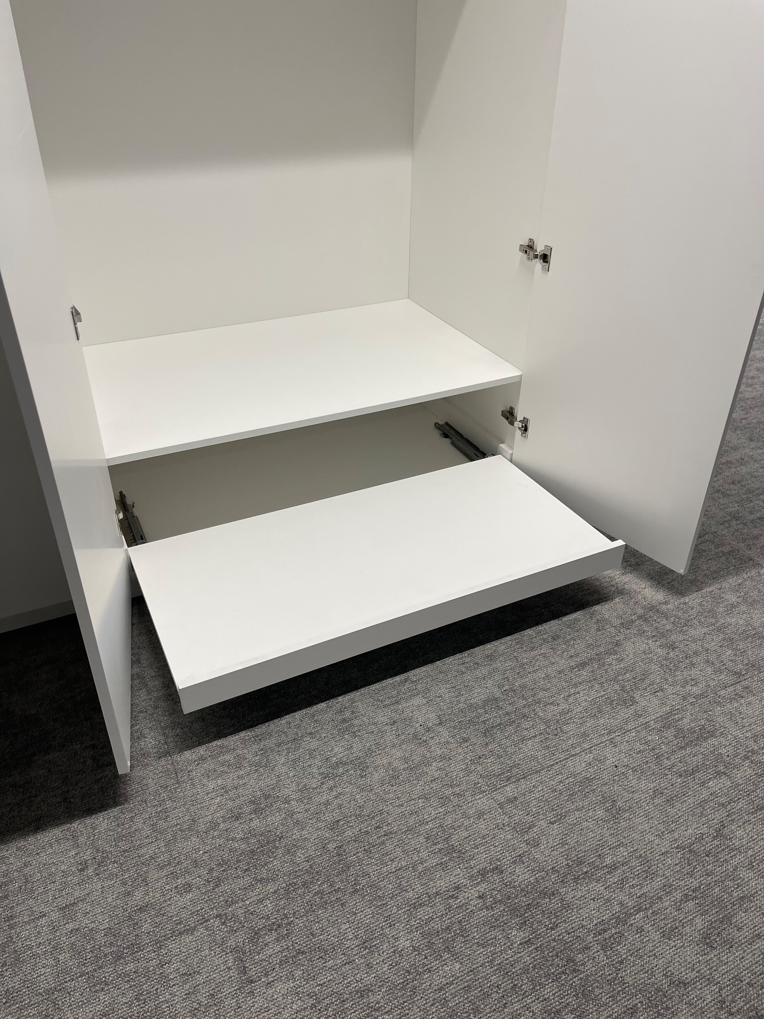 Wnętrze białej szafy z wysuwaną półką na metalowych prowadnicach, widoczne zawiasy drzwi. Na podłodze szara wykładzina dywanowa.