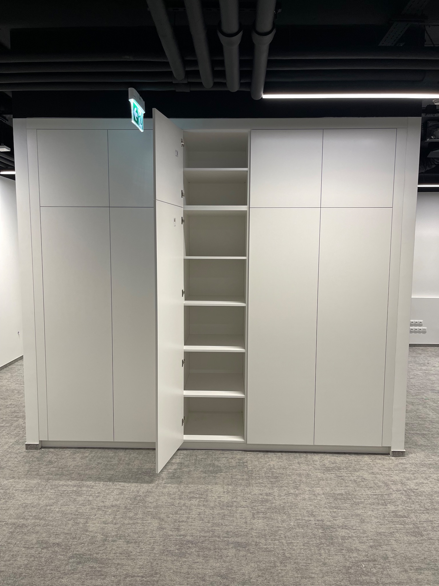 Biała szafa wnękowa z otwartymi drzwiami, widoczne półki. Minimalistyczny design, szara wykładzina podłogowa, sufit z instalacjami. Oświetlenie biurowe.