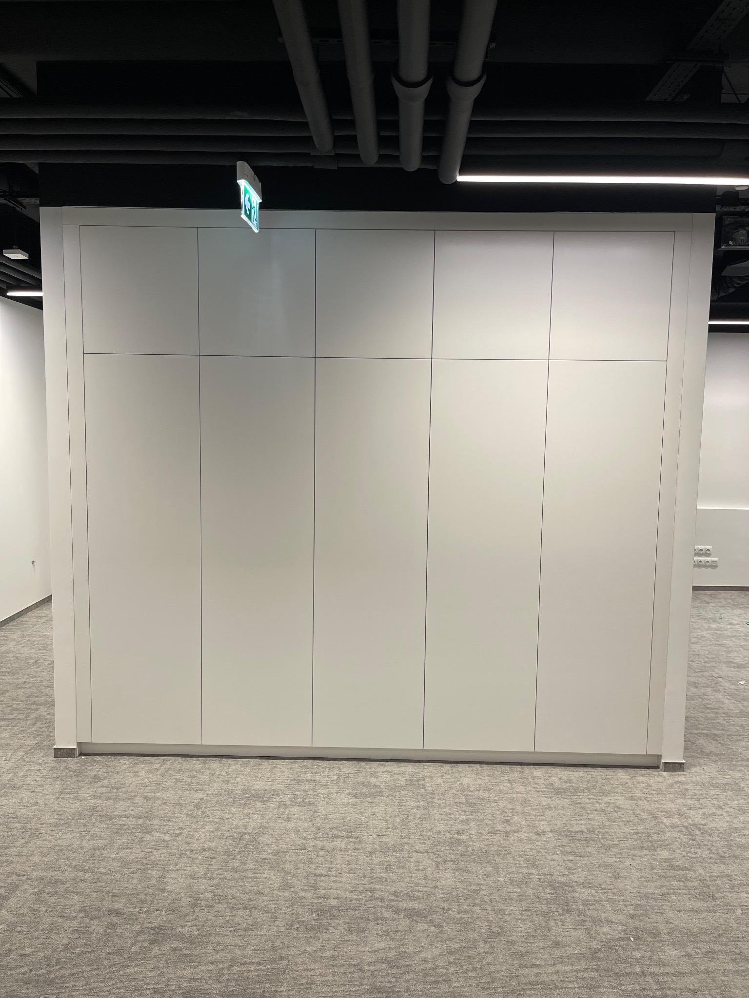 Biała, minimalistyczna szafa wnękowa z sześcioma drzwiami, umieszczona w biurowym wnętrzu z szarą wykładziną i widocznymi instalacjami pod sufitem.