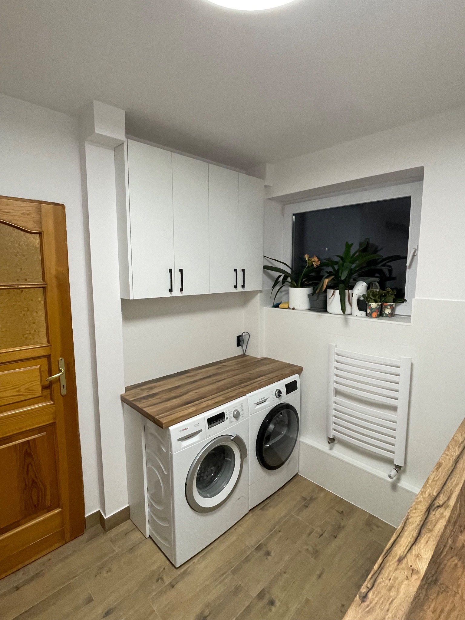 Białe szafki wiszące z czarnymi uchwytami nad pralką i suszarką z drewnianym blatem. Na parapecie rośliny doniczkowe. Drewniane drzwi po lewej.