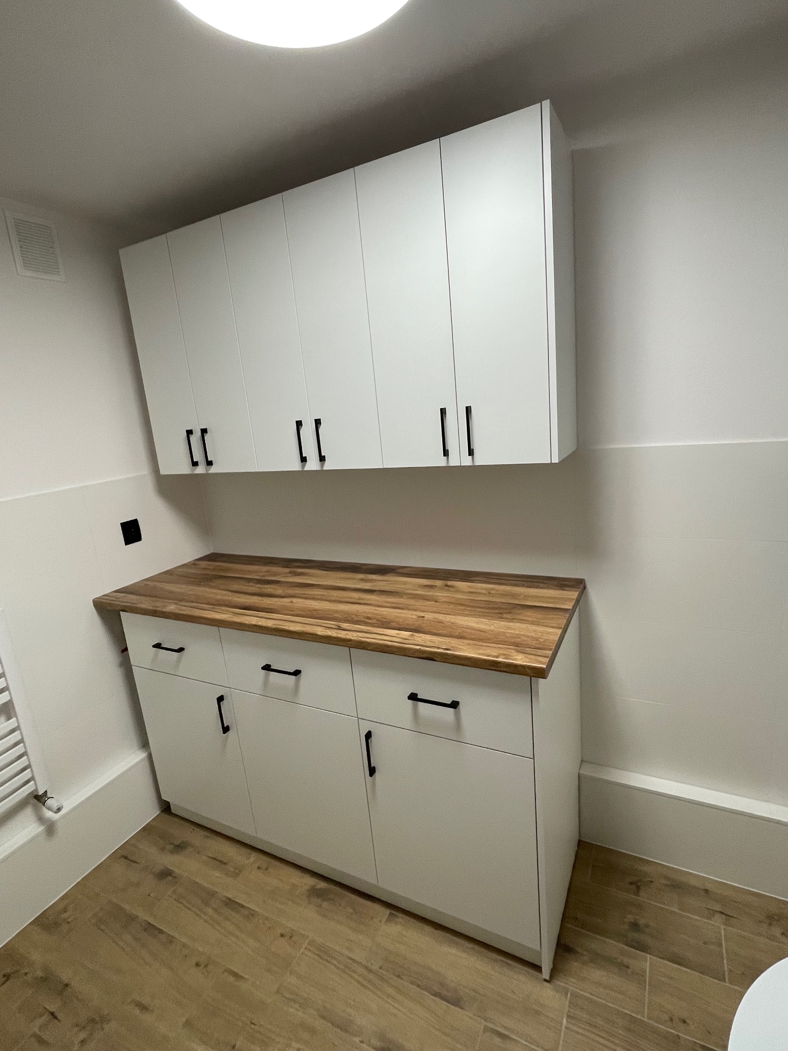 Białe szafki łazienkowe z drewnianym blatem i czarnymi uchwytami, wiszące nad podłogą z płytek imitujących drewno. Minimalistyczny design, jasne wnętrze.