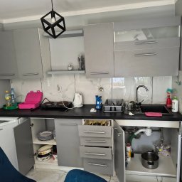 Domowa Harmonia - Kadr kuchni w Przemęcie przed sprzątaniem: szare szafki, otwarte drzwiczki, naczynia, bałagan na blacie i pod zlewem. Ujęcie z dołu, widoczny fragment krzesła.