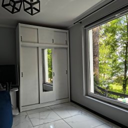 Domowa Harmonia - Jasne wnętrze pokoju po remoncie: biała szafa z lustrem, okno z widokiem na zieleń, marmurowe płytki podłogowe i nowoczesne lampy.