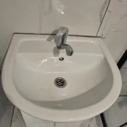 Biała umywalka z chromowaną baterią na tle marmurowych płytek. Widoczny fragment szarej wycieraczki i kabiny prysznicowej. Ujęcie z g&oacute;ry.