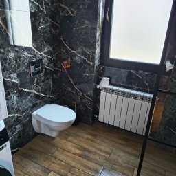 Domowa Harmonia - Elegancka łazienka po remoncie: czarne marmurowe ściany z białymi żyłami, drewnopodobna podłoga, biała toaleta podwieszana i grzejnik.
