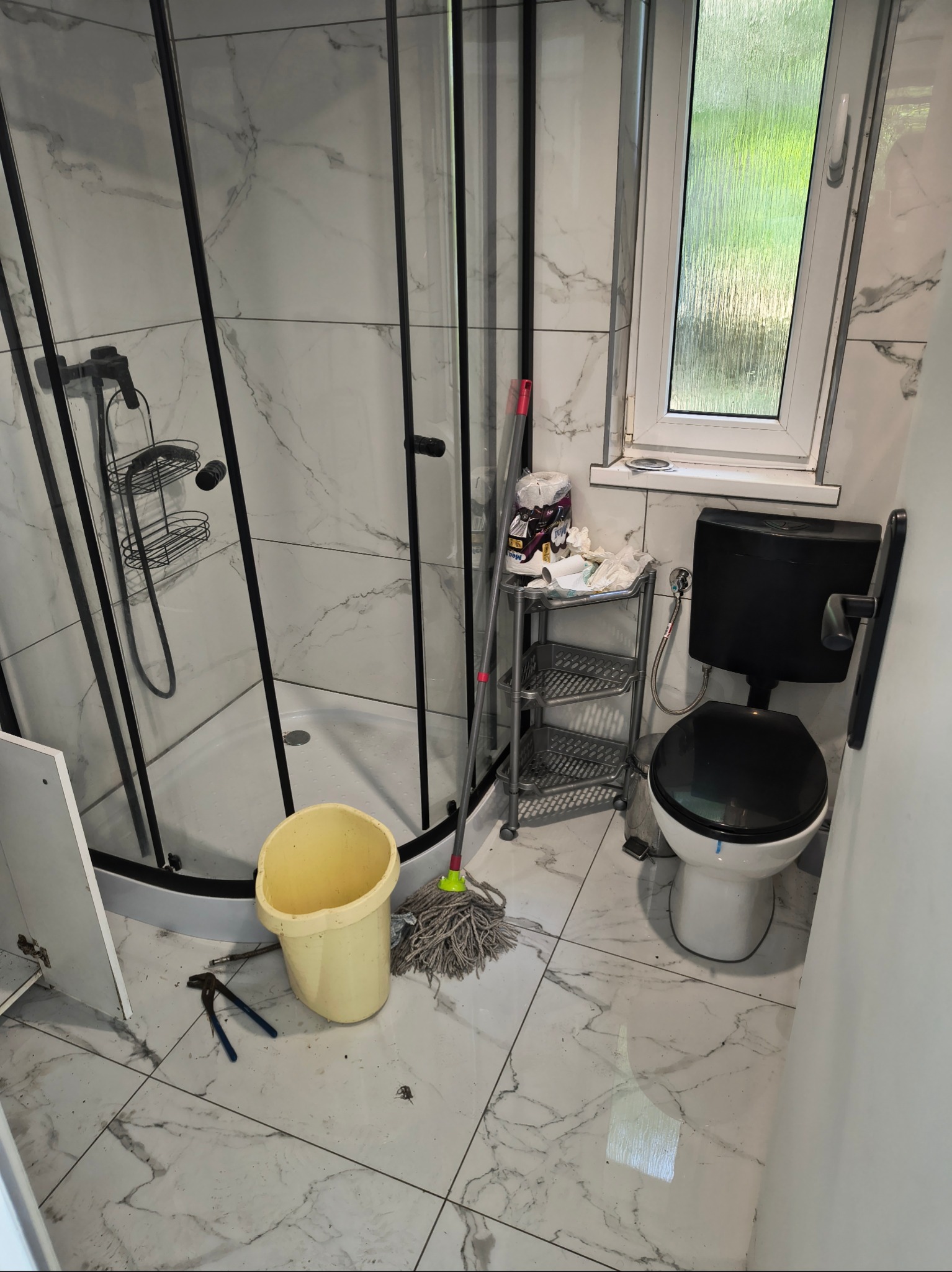 Łazienka przed sprzątaniem: widoczna kabina prysznicowa, toaleta, mop i wiadro. Na podłodze widoczne zabrudzenia i narzędzia. Marmurowe płytki na ścianach i podłodze.