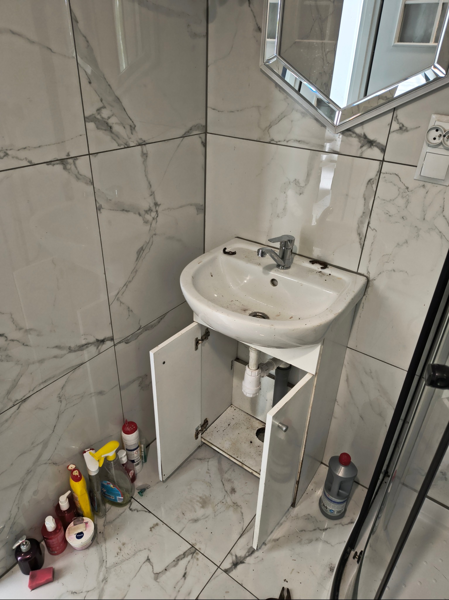 Zabrudzona umywalka z otwartą szafką w łazience wyłożonej marmurowymi płytkami. Widoczne środki czystości i lustro z geometryczną ramą.