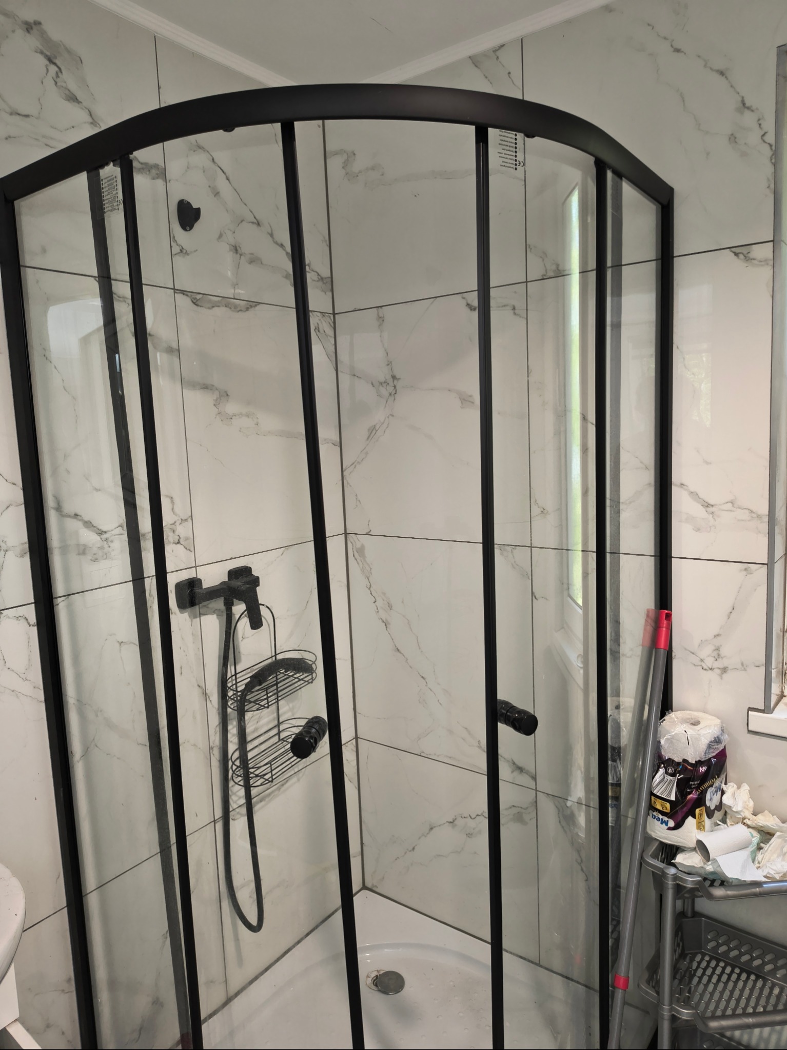 Narożna kabina prysznicowa z czarnymi profilami na tle marmurowych płytek. Widoczne akcesoria i niedoskonałości przed sprzątaniem.