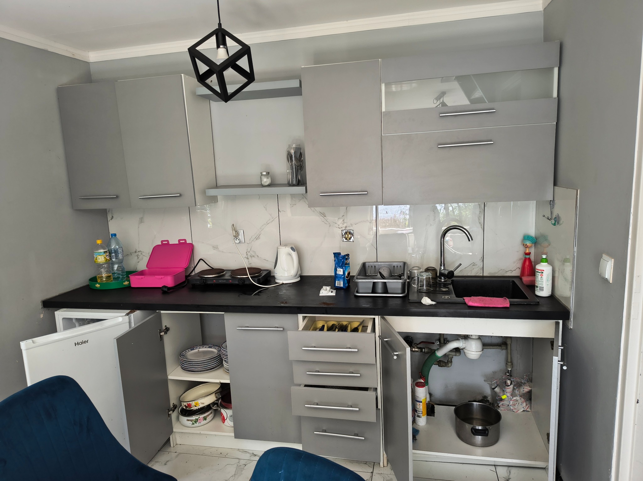 Kadr kuchni w Przemęcie przed sprzątaniem: szare szafki, otwarte drzwiczki, naczynia, bałagan na blacie i pod zlewem. Ujęcie z dołu, widoczny fragment krzesła.