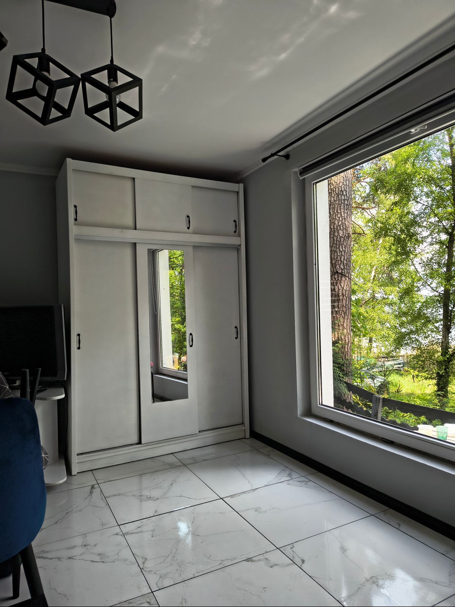 Jasne wnętrze pokoju po remoncie: biała szafa z lustrem, okno z widokiem na zieleń, marmurowe płytki podłogowe i nowoczesne lampy.