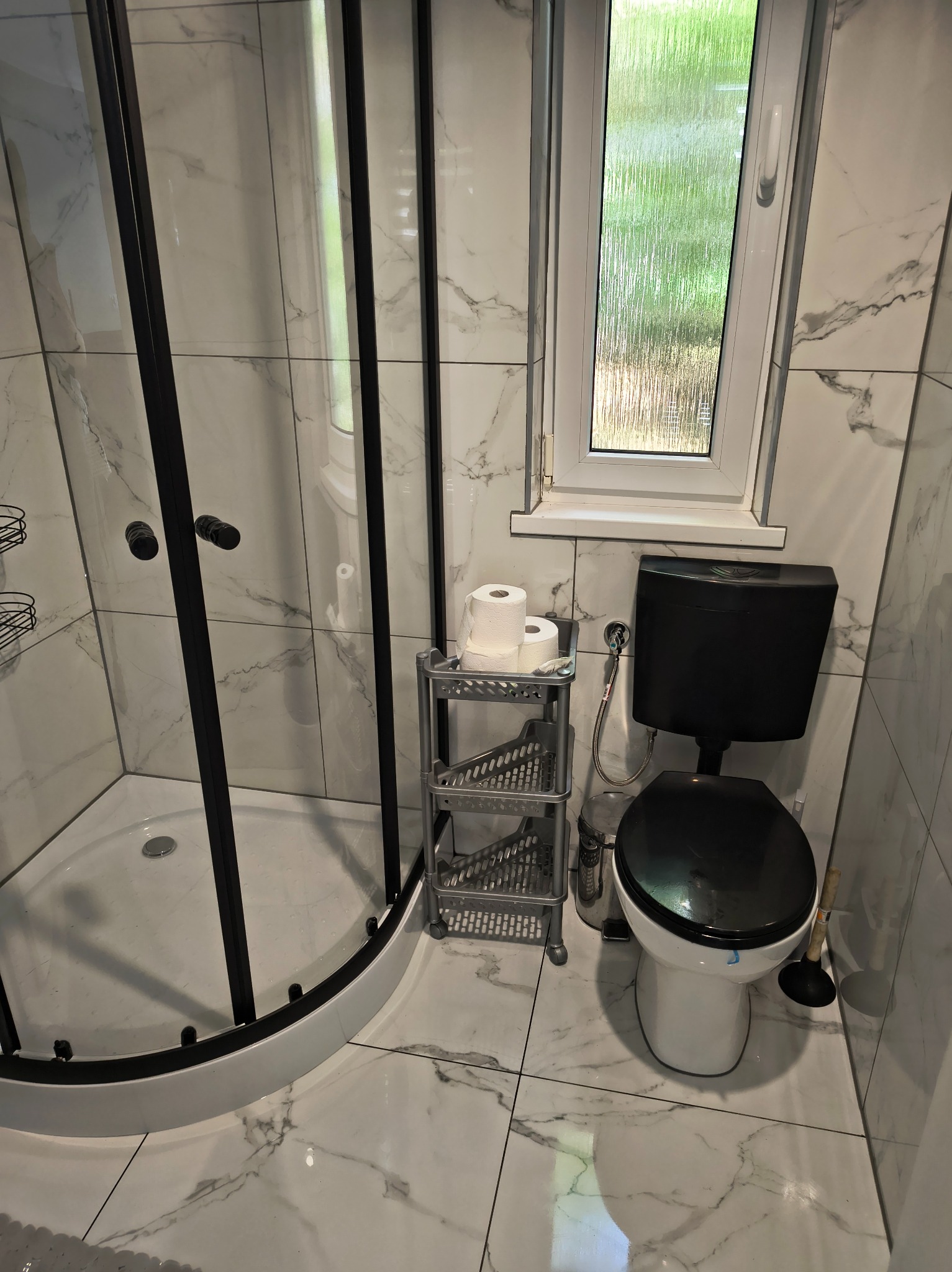 Nowoczesna łazienka po remoncie: kabina prysznicowa z czarnymi ramami, toaleta w czarno-białej kolorystyce, marmurowe płytki i okno z widokiem na zieleń.