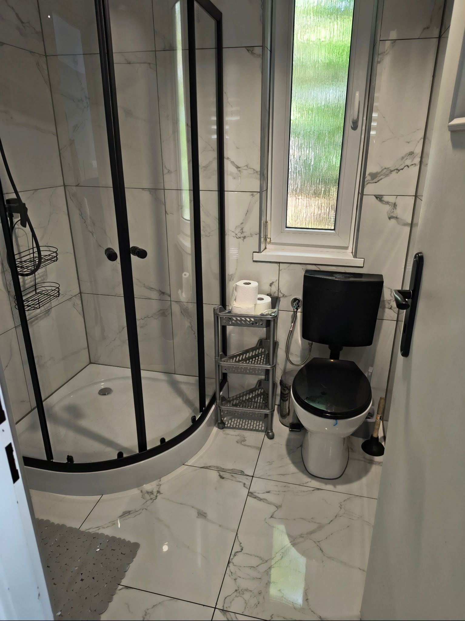 Wykończona łazienka z kabiną prysznicową z czarnymi profilami, toaletą i marmurowymi płytkami. Widok z perspektywy wejścia do pomieszczenia.