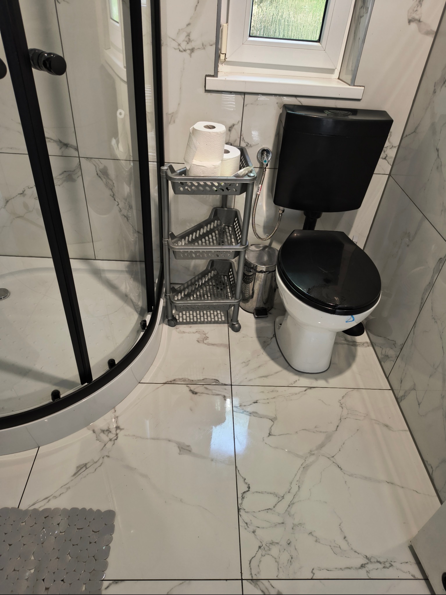 Wykończona łazienka z kabiną prysznicową, toaletą z czarną spłuczką i białym sedesem oraz marmurowymi płytkami. Widoczny regał z papierem toaletowym.