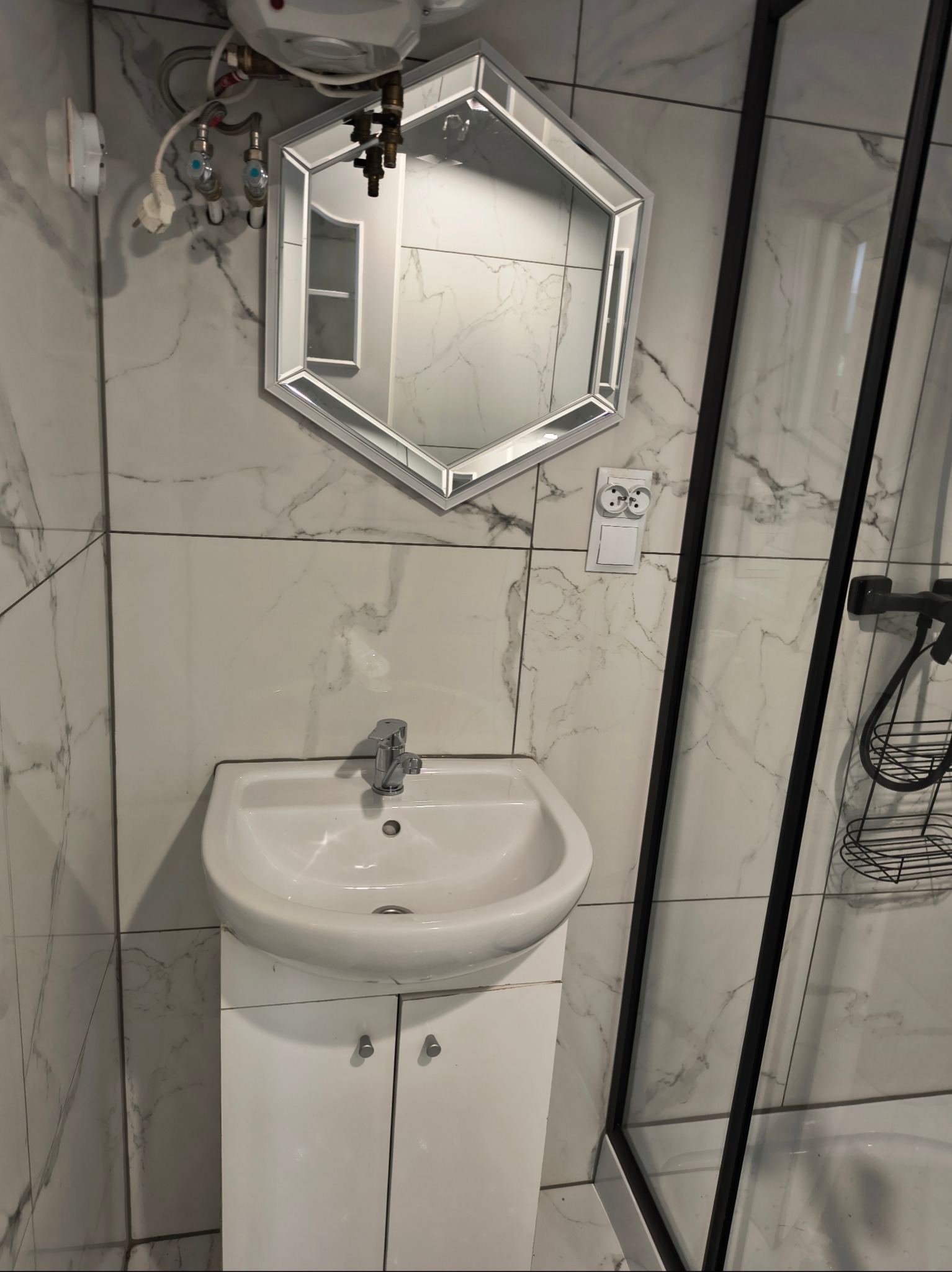 Biała umywalka z szafką w nowoczesnej łazience z marmurowymi płytkami, lustrem w kształcie heksagonu i czarną kabiną prysznicową.