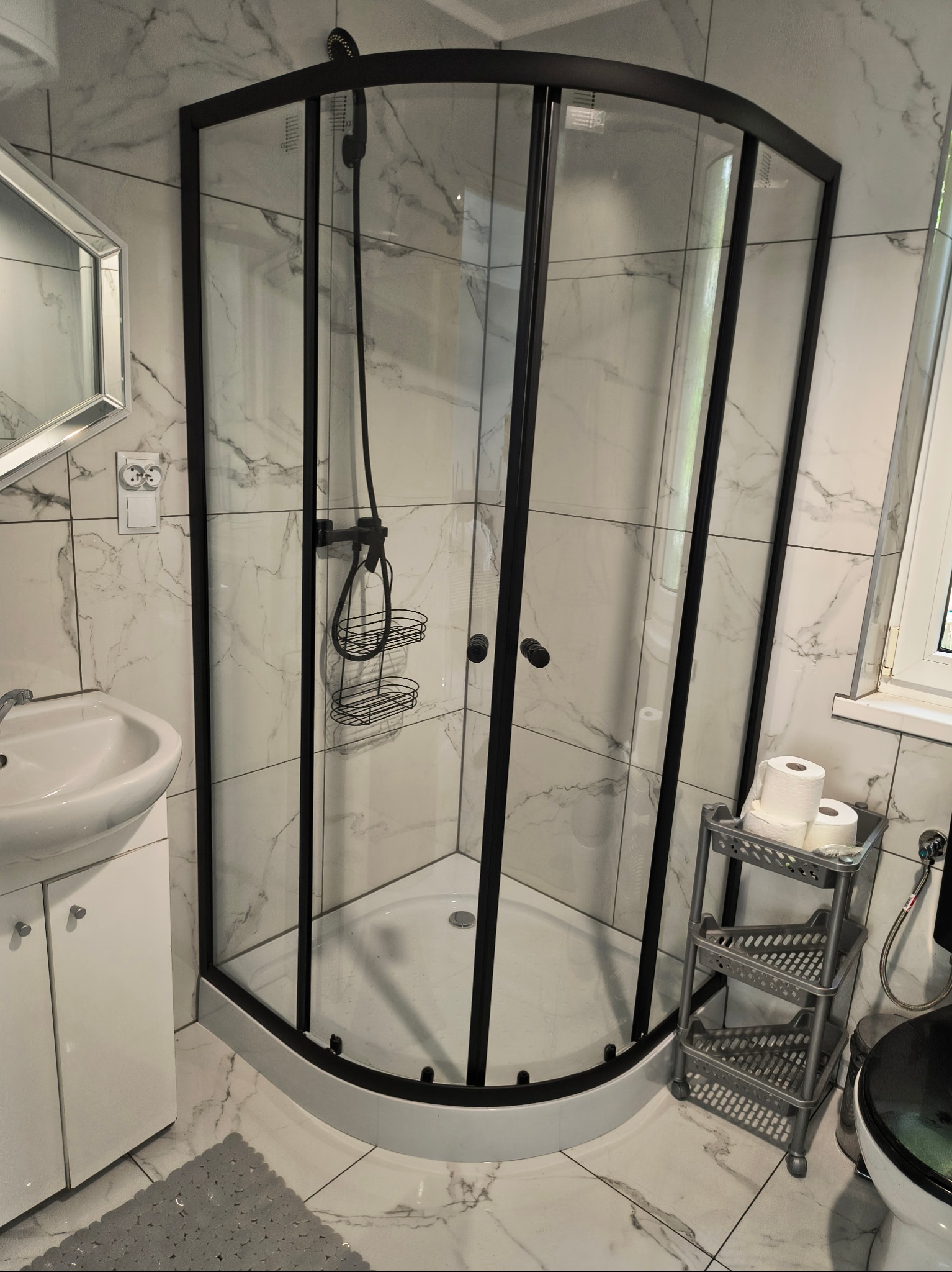 Nowoczesna kabina prysznicowa z czarnymi ramami i marmurowymi płytkami w łazience z białą umywalką i akcesoriami. Widok z góry.