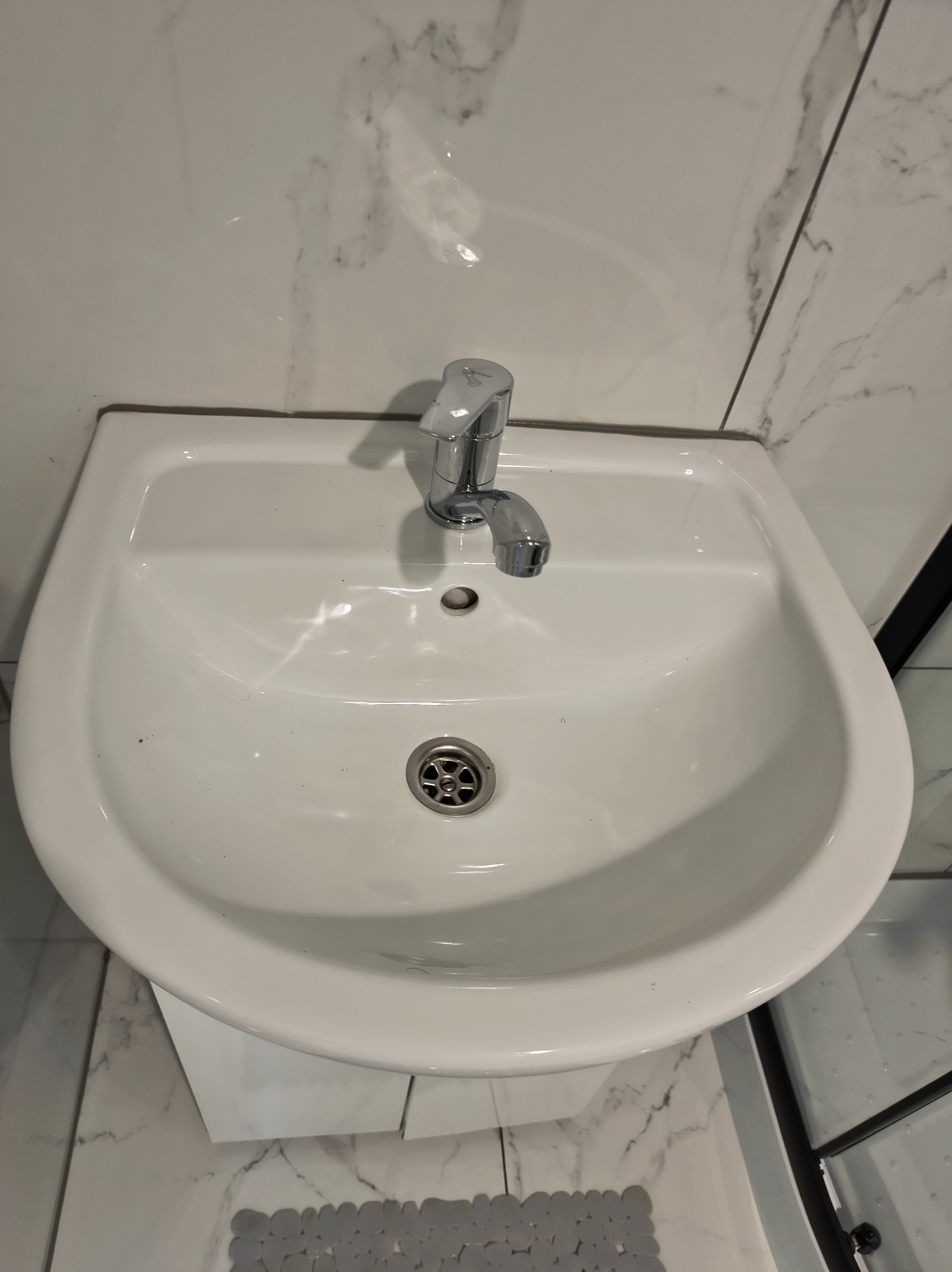 Biała umywalka z chromowaną baterią na tle marmurowych płytek. Widoczny fragment szarej wycieraczki i kabiny prysznicowej. Ujęcie z góry.