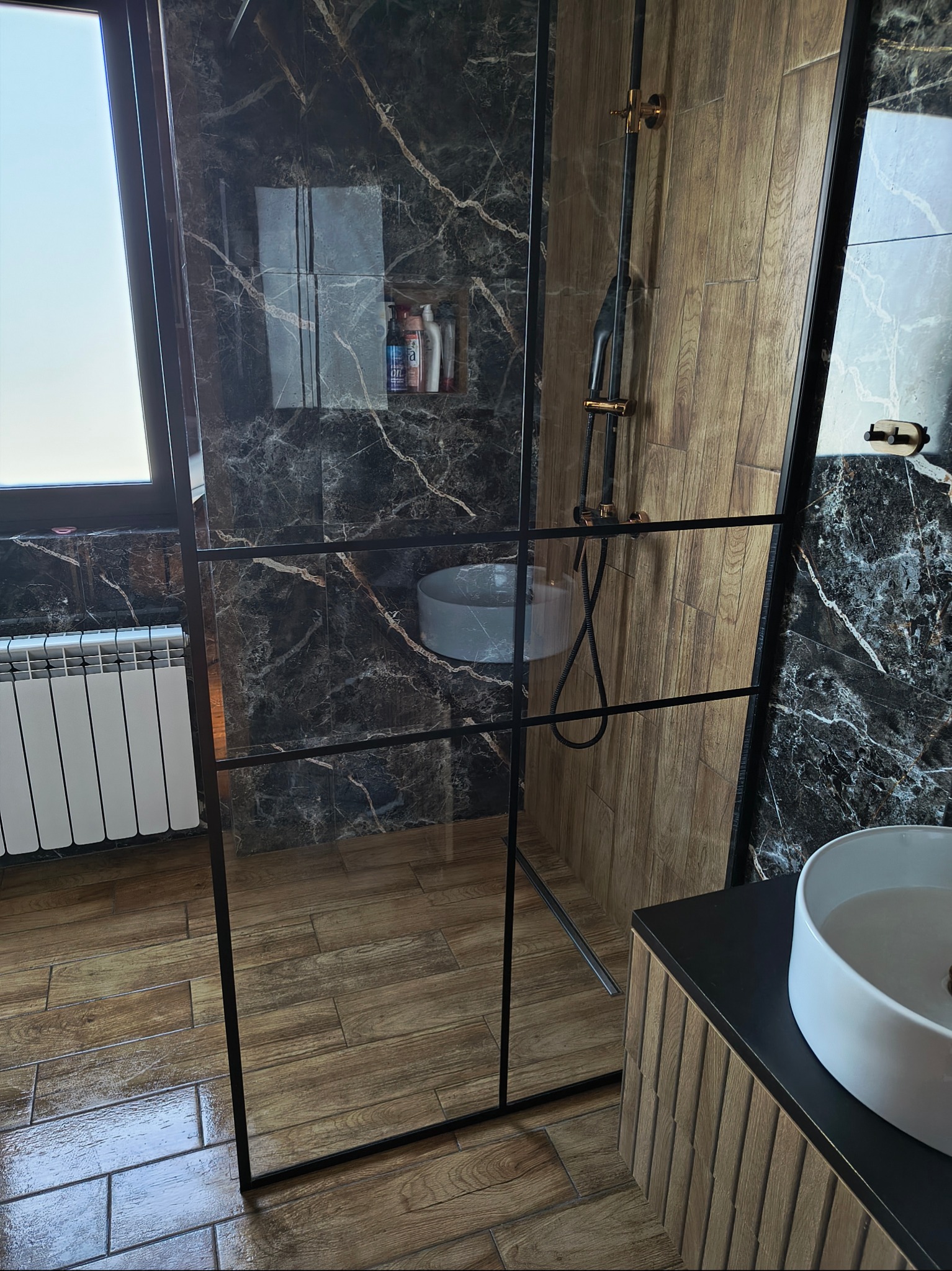 Nowoczesna kabina prysznicowa typu walk-in z czarnymi profilami, wykończona marmurem i drewnem. Widoczna armatura w kolorze złota i okno z grzejnikiem.