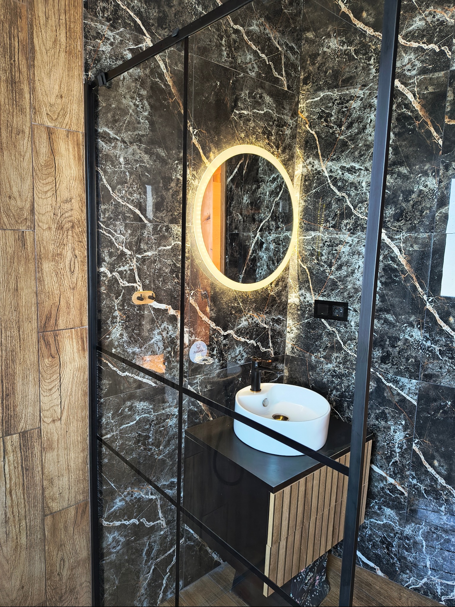 Nowoczesna łazienka po remoncie: czarne marmurowe płytki, okrągłe lustro z podświetleniem, biała umywalka na szafce z drewnianymi lamelami.