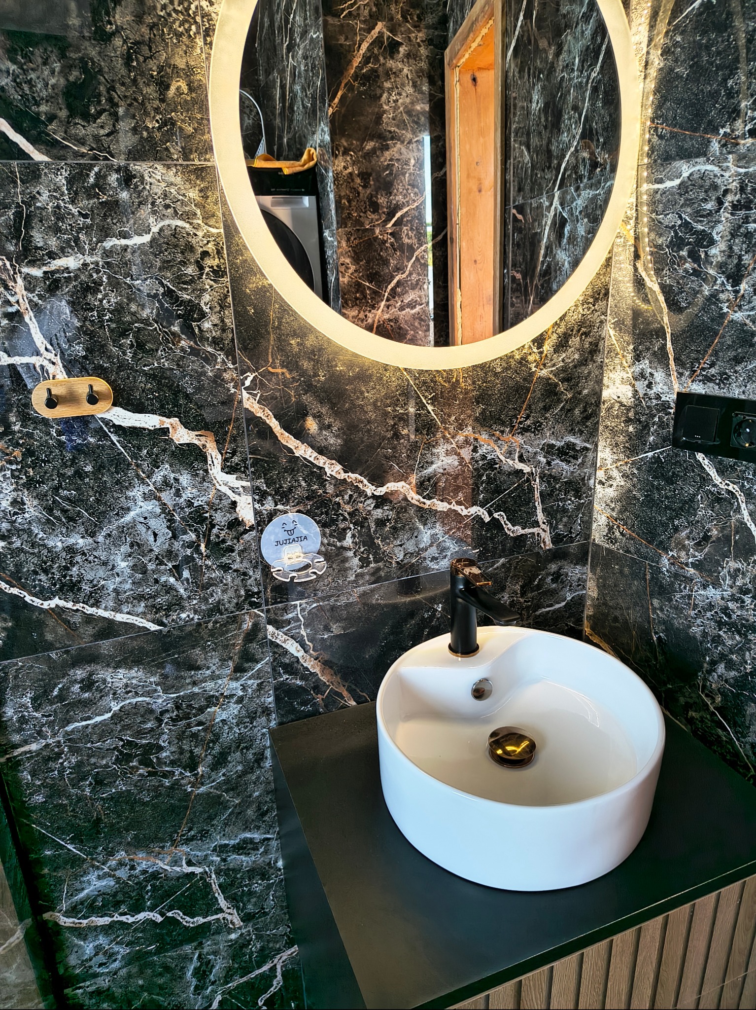 Nowoczesna łazienka z umywalką nablatową i okrągłym lustrem z podświetleniem LED na ścianie z czarnego marmuru z złotymi żyłami.