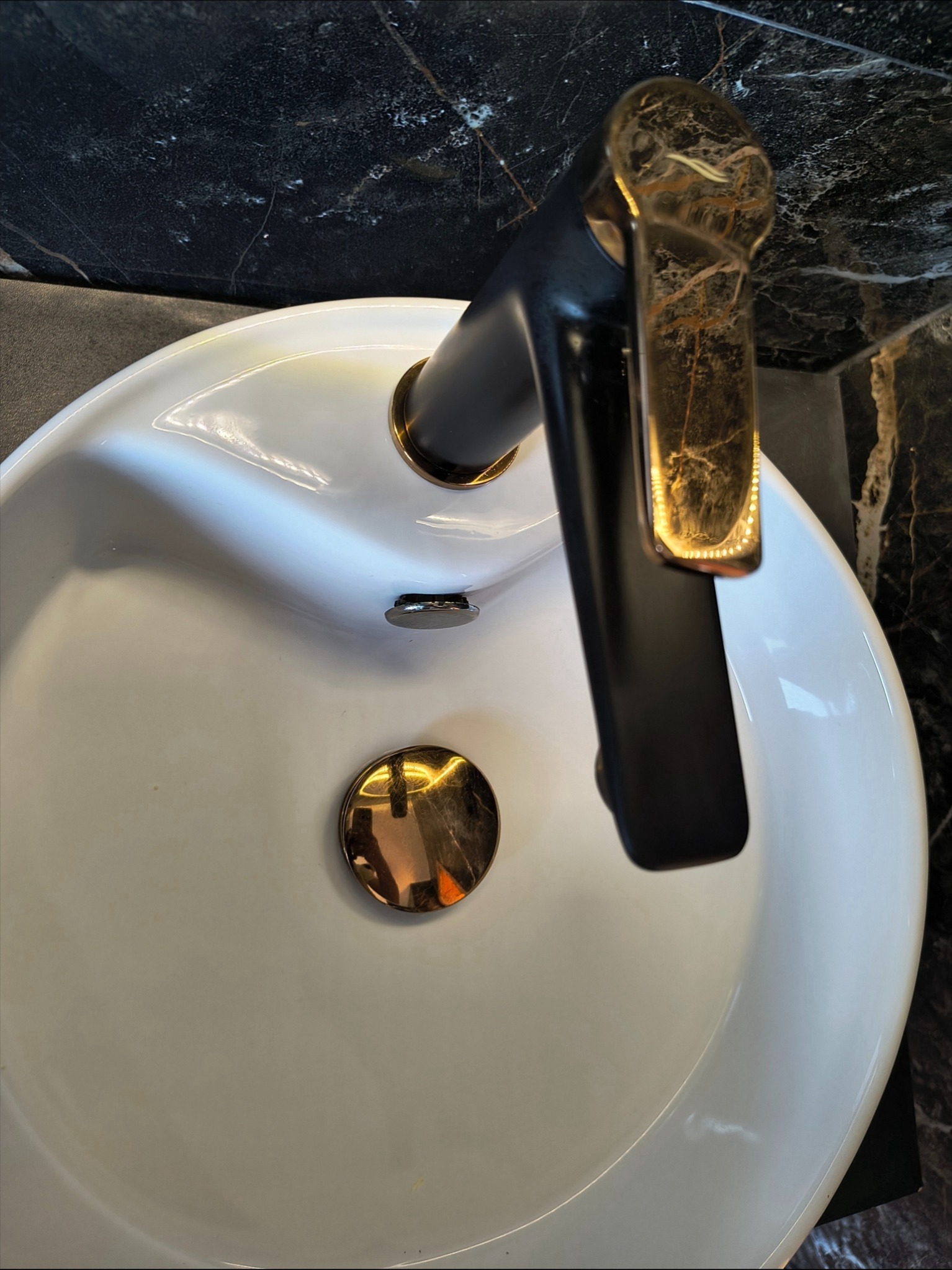 Biała umywalka z czarno-złotą baterią na tle czarnego marmuru. Bateria ma unikalny wzór, przypominający żyły marmuru. Widok z góry.