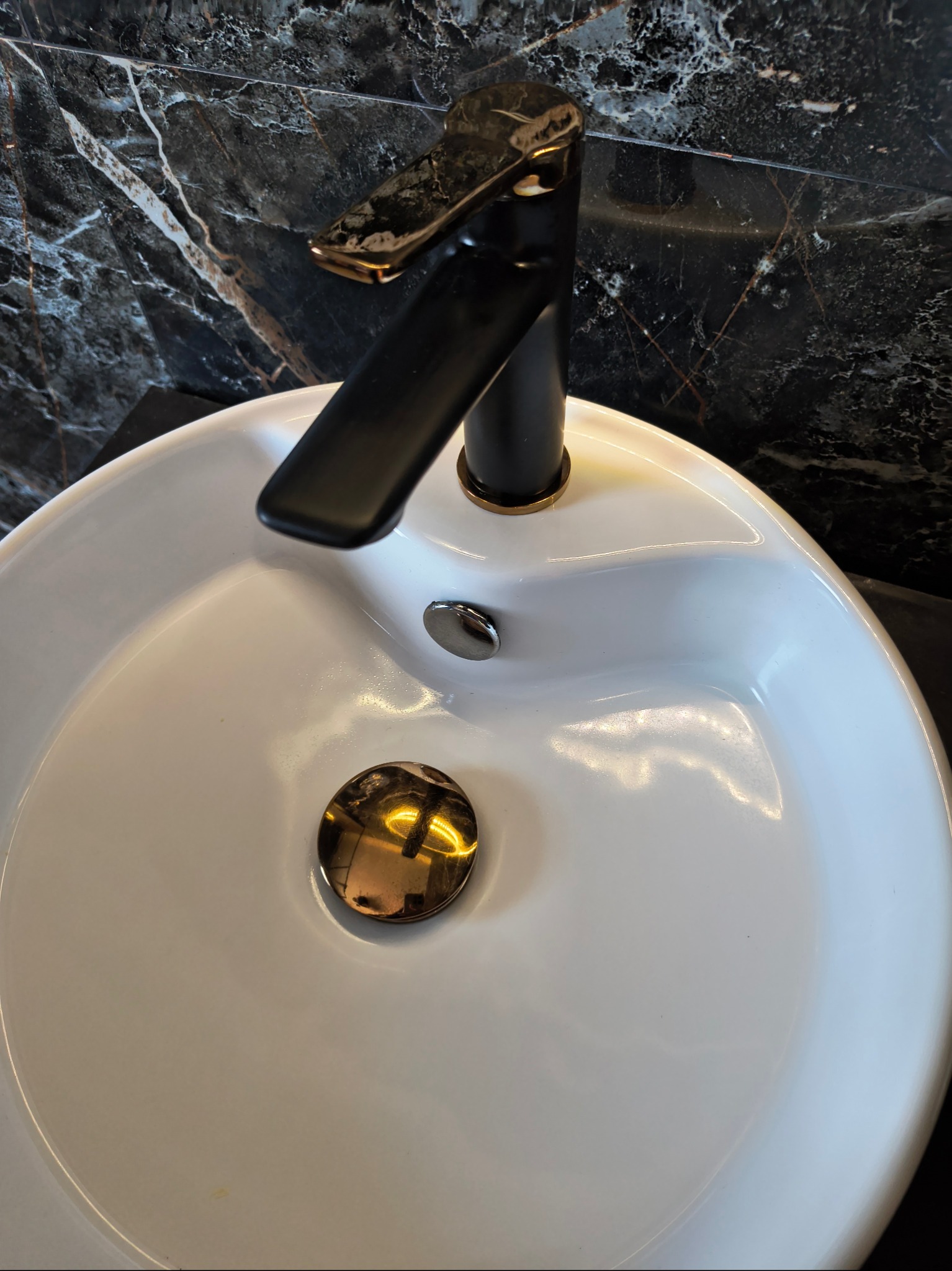 Biała umywalka z czarno-złotą armaturą na tle czarnego marmuru. Błyszcząca powierzchnia, nowoczesny design, widoczne odbicia światła.