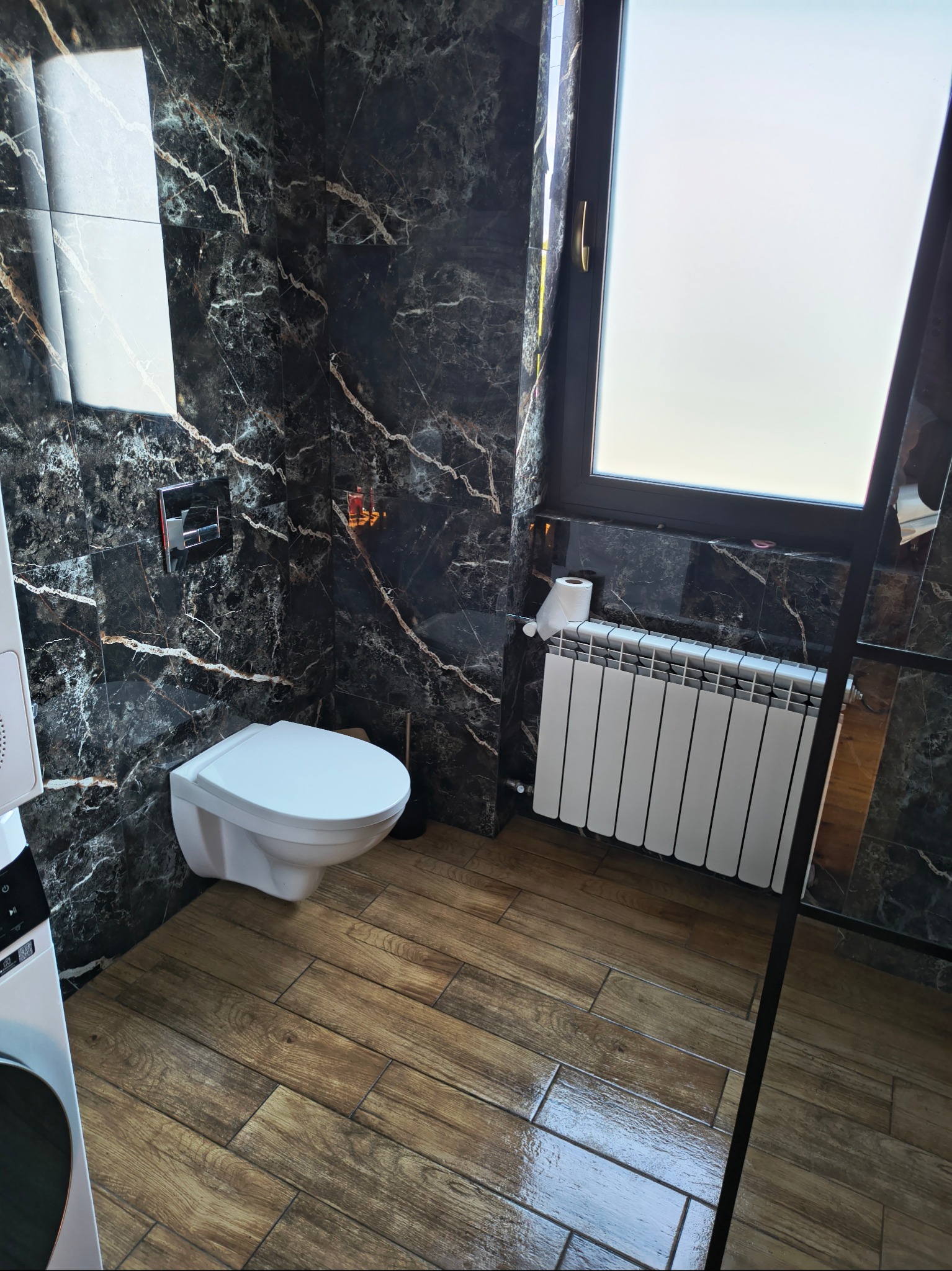 Elegancka łazienka po remoncie: czarne marmurowe ściany z białymi żyłami, drewnopodobna podłoga, biała toaleta podwieszana i grzejnik.