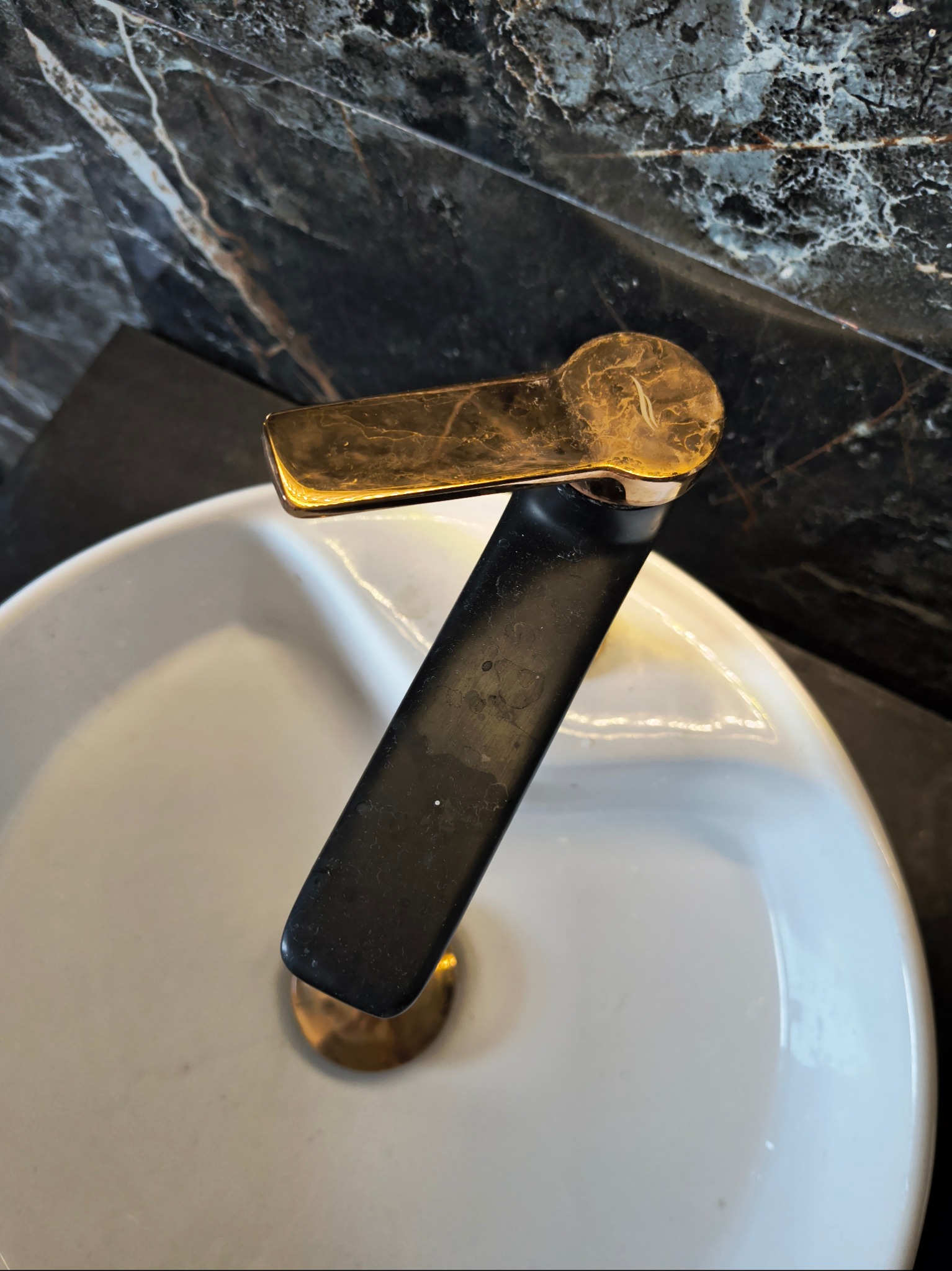 Bateria umywalkowa z czarnym korpusem i złotą rączką, widoczna na tle marmurowej ściany i białej umywalki. Zdjęcie z bliska, ukazujące detale.