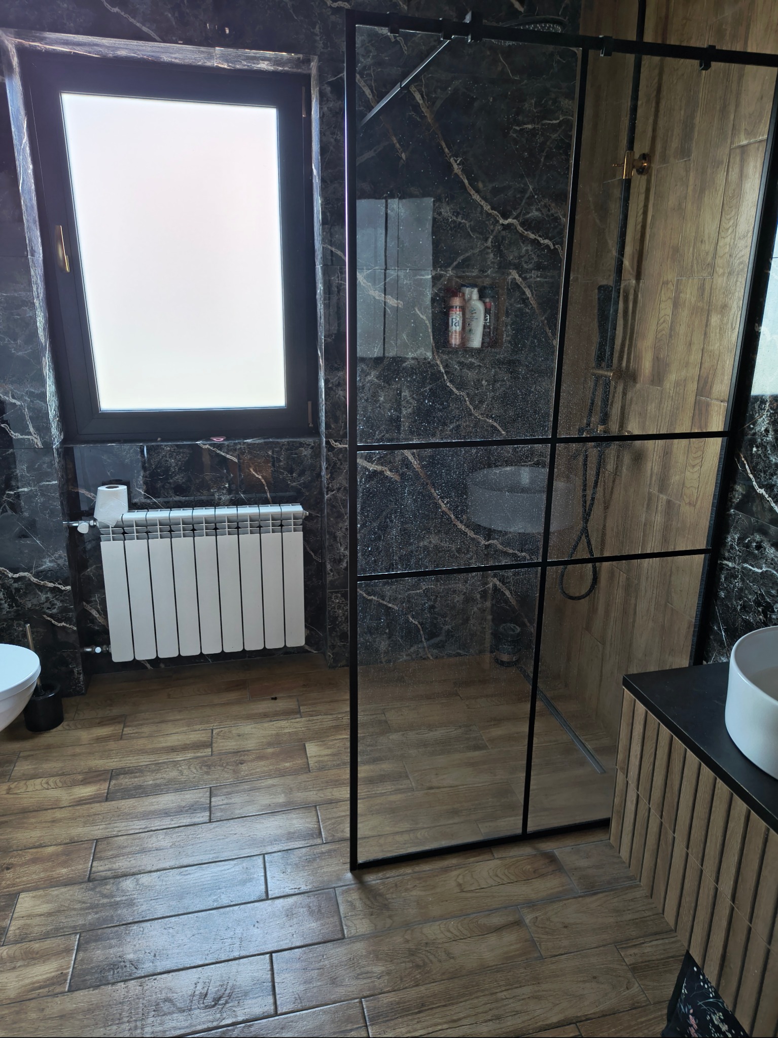 Ciemna łazienka przed sprzątaniem: marmurowe ściany, drewniana podłoga, kabina prysznicowa z czarnymi ramami, okno z białym grzejnikiem. Widok na umywalkę.
