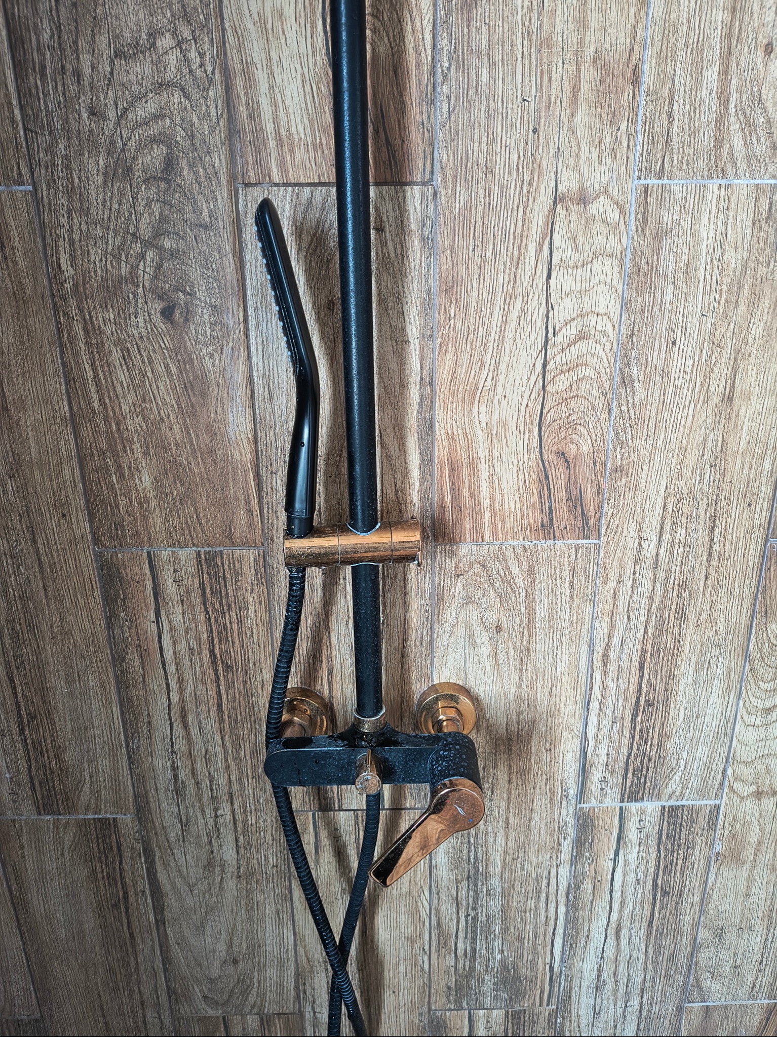 Czarno-złota bateria prysznicowa na drewnopodobnej ścianie, widok z przodu. Element łazienki w stylu retro.