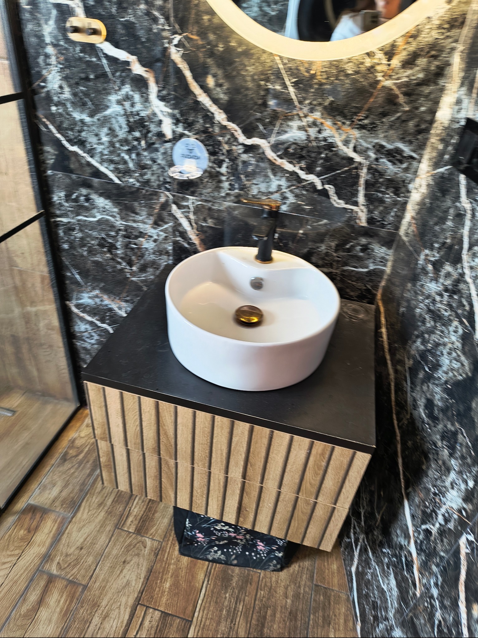 Nowoczesna umywalka nablatowa w łazience z marmurową ścianą i drewnianą szafką. Czarna bateria i okrągłe lustro z podświetleniem. Widok z góry na podłogę.