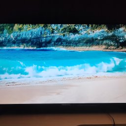 Piksel 101 - Włączony telewizor Manta wyświetla krystalicznie czysty obraz plaży z turkusową wodą i klifami. Widoczne detale i żywe kolory.