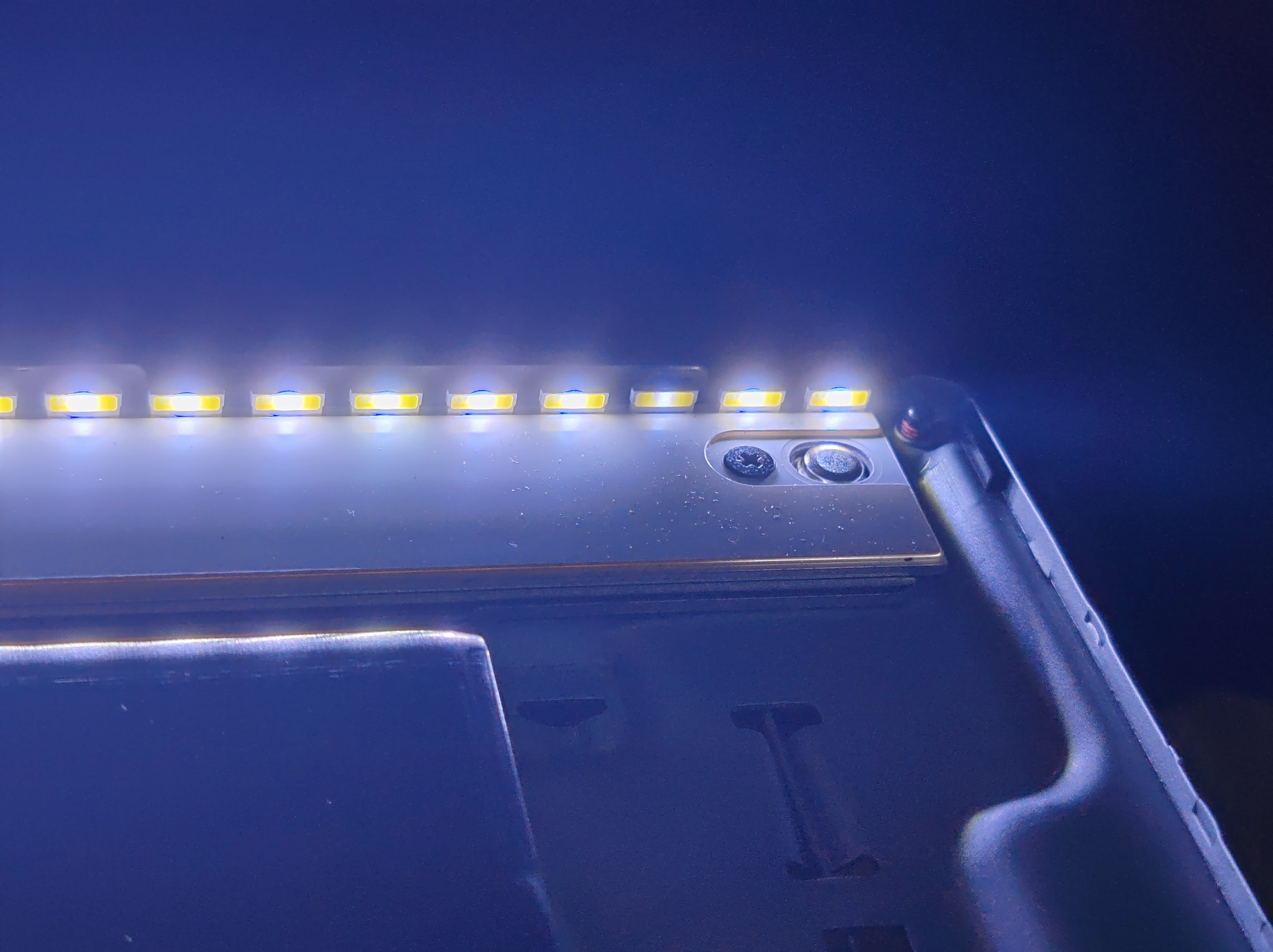 Otwarta obudowa telewizora z widoczną listwą LED podświetlającą matrycę. Widoczne ślady naprawy i demontażu. Niebieskie tło.