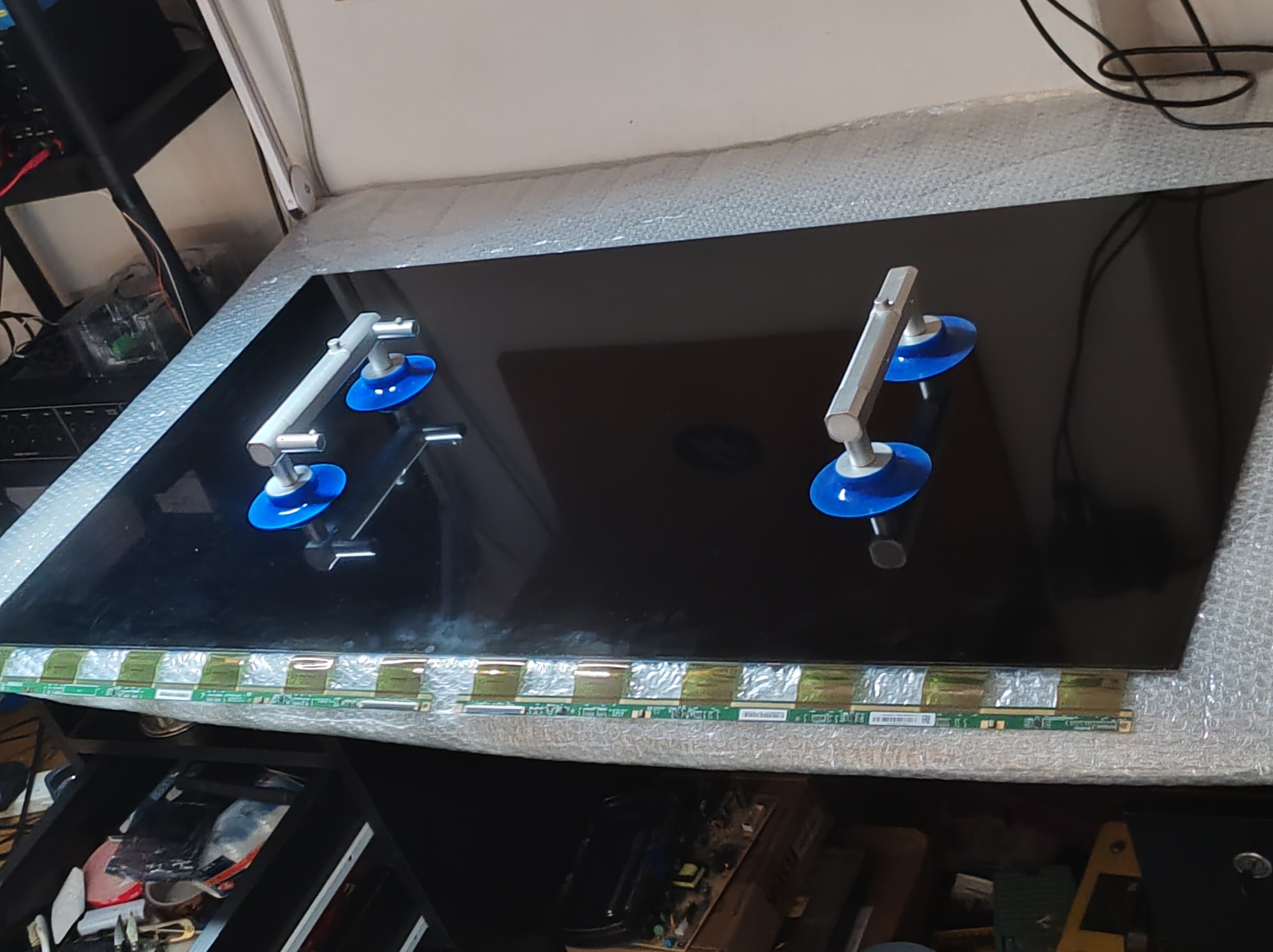 Ekran telewizora zabezpieczony przyssawkami podczas naprawy. Widoczne taśmy połączeniowe i narzędzia w tle. Blat roboczy z fakturą.