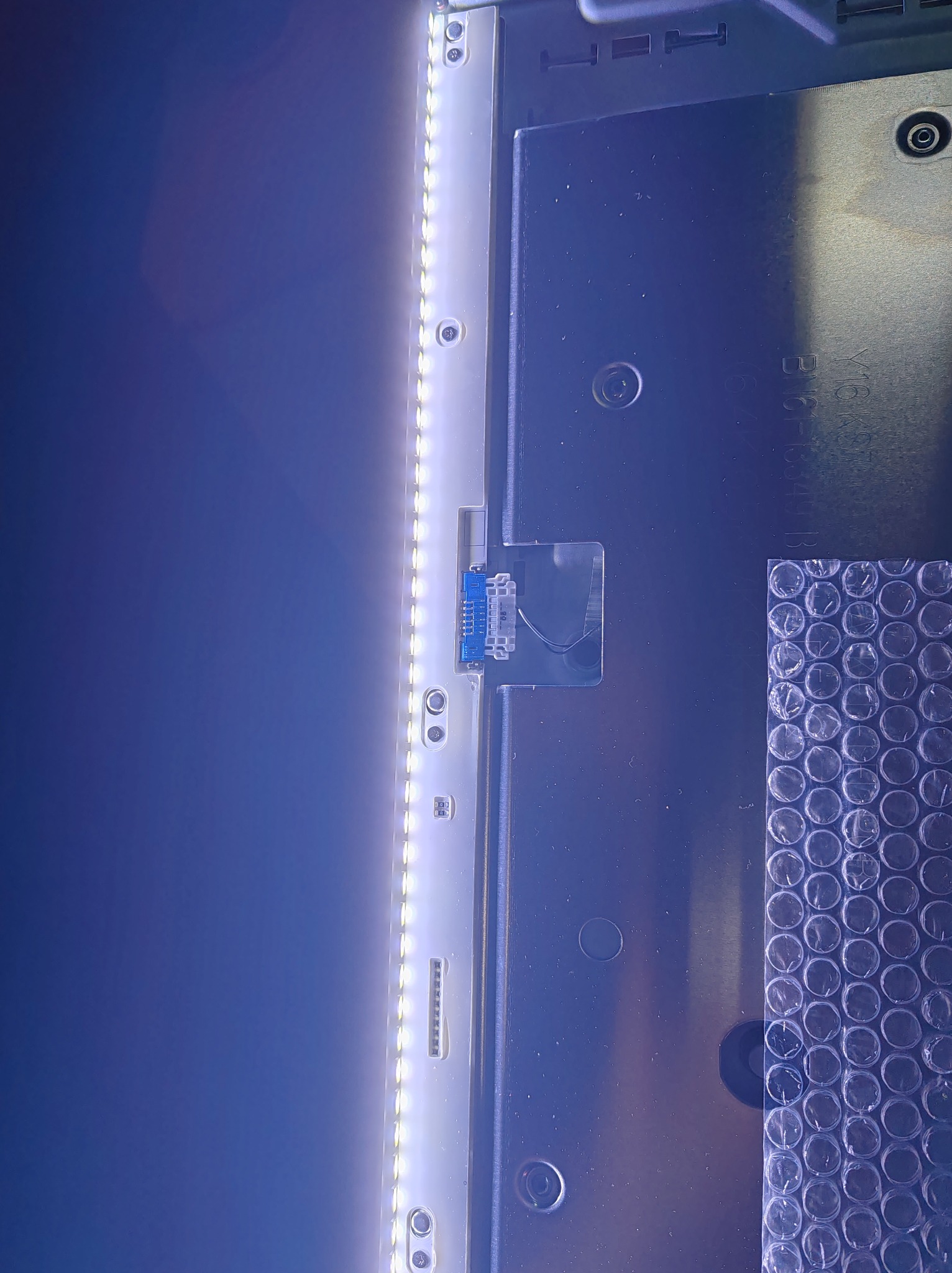 Wnętrze telewizora z podświetleniem LED, widoczne złącze i fragment folii bąbelkowej. Detal z naprawy sprzętu RTV, prezentujący elementy techniczne.