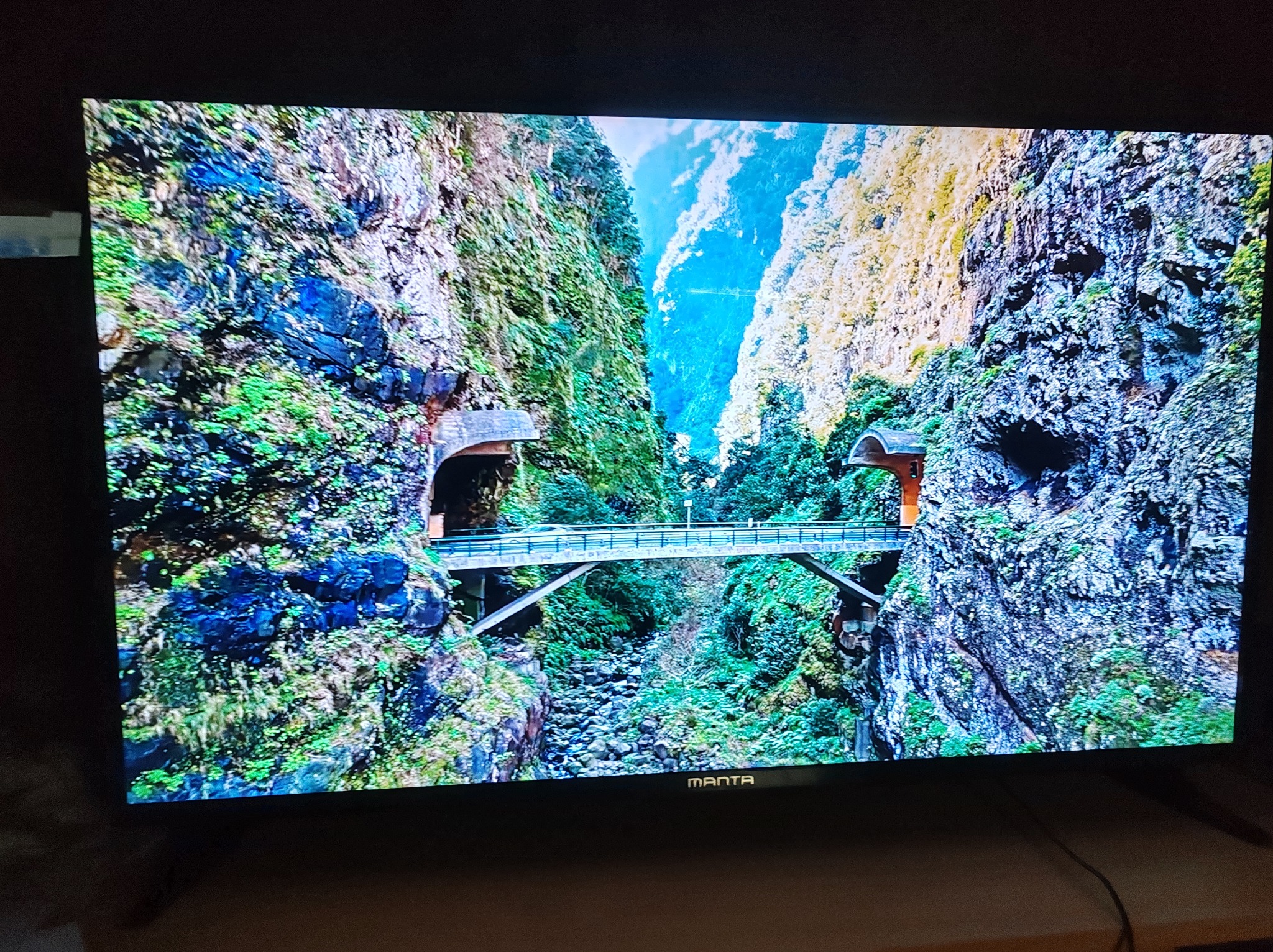 Włączony telewizor Manta wyświetla krajobraz górski z mostem. Widoczne detale skał i roślinności, oświetlenie naturalne. Kabel zasilający widoczny w dolnej części.