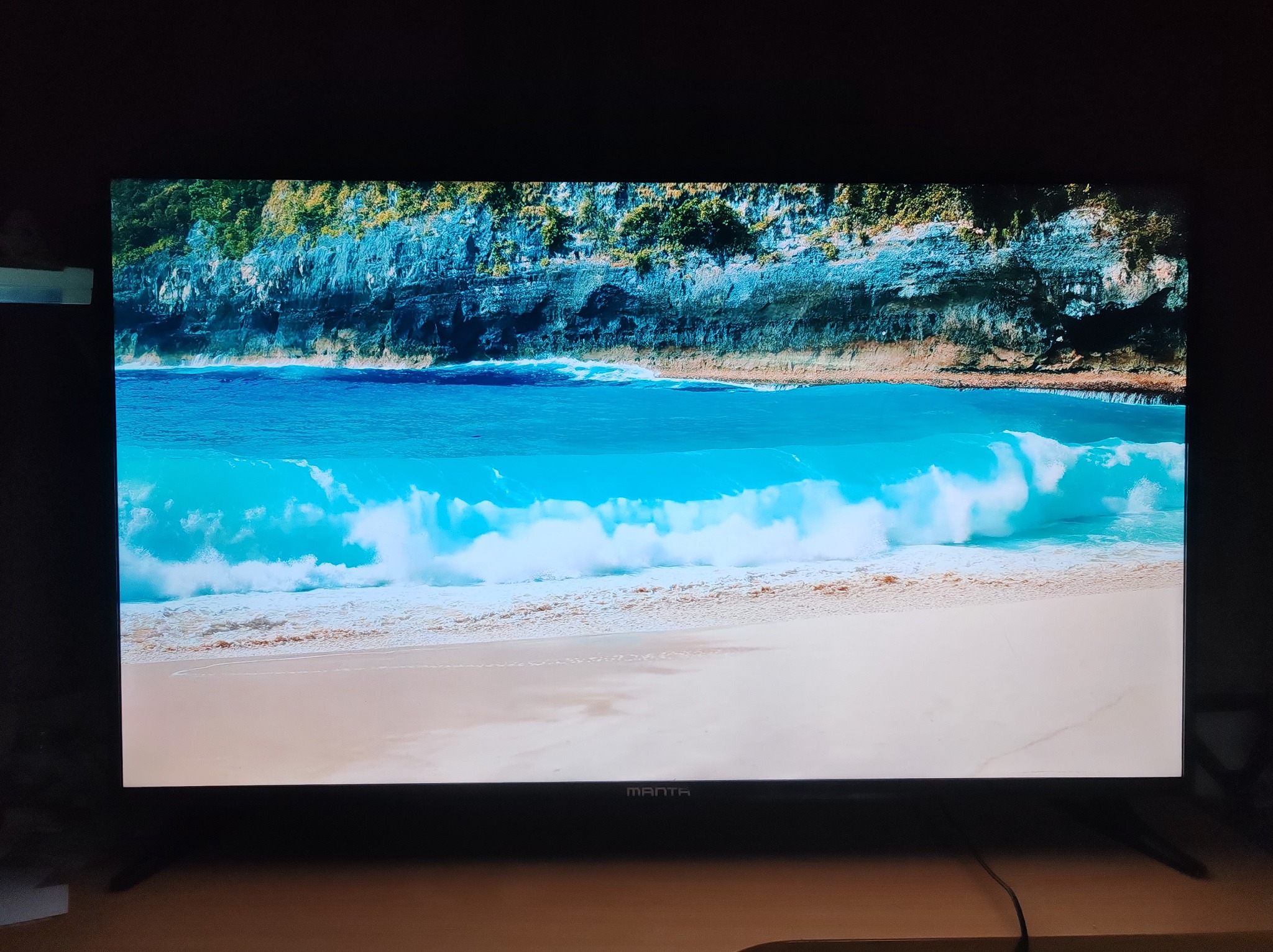 Włączony telewizor Manta wyświetla krystalicznie czysty obraz plaży z turkusową wodą i klifami. Widoczne detale i żywe kolory.