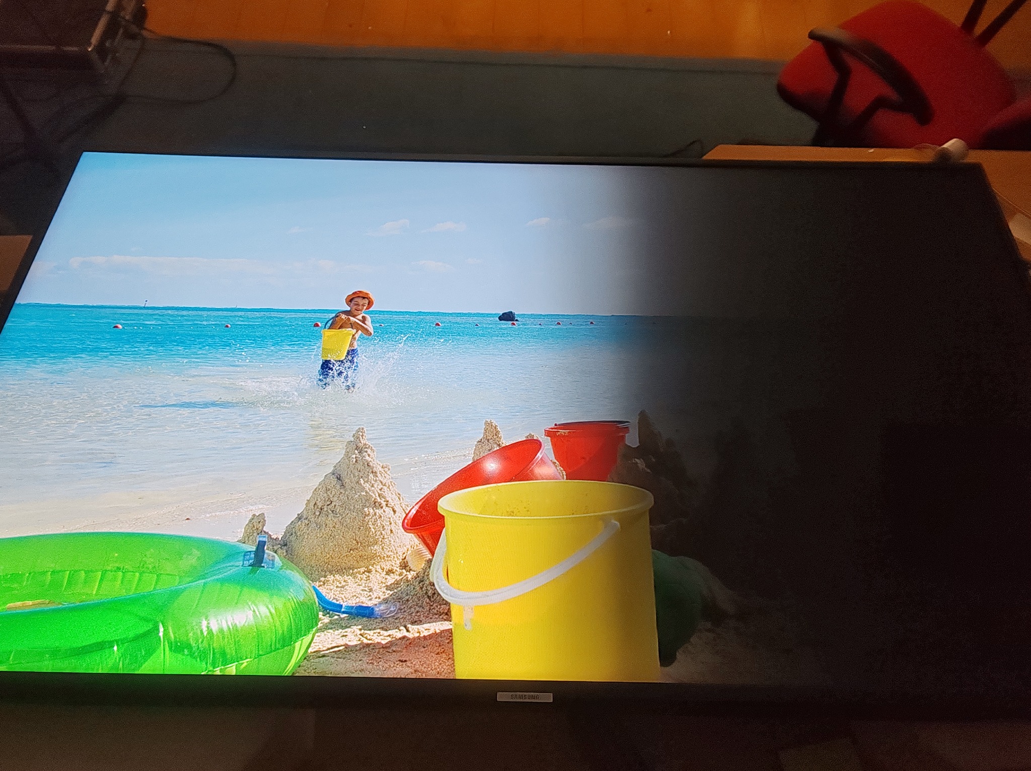 Wyłączony telewizor LCD, na którego części ekranu widoczna jest scena z plaży z dzieckiem i zabawkami. Prawa strona ekranu jest ciemna.