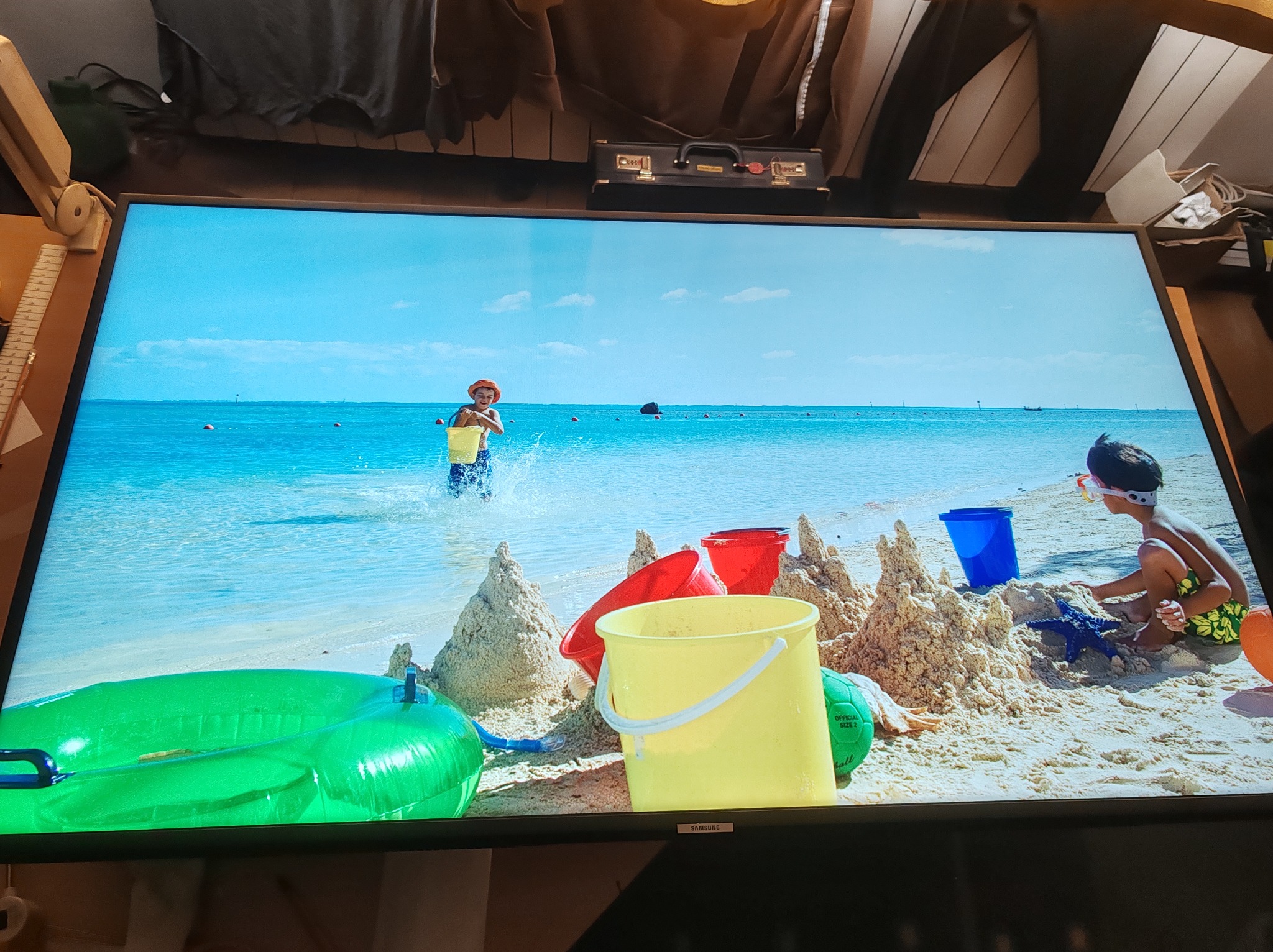 Włączony telewizor Samsung wyświetla plażę z dziećmi, piaskowe zamki, wiaderka i dmuchane koło. Nad telewizorem widoczny kufer i ubrania.