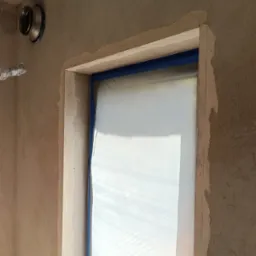 Ściana w trakcie szpachlowania przy oknie, zabezpieczonym folią i taśmą. Widoczny fragment instalacji wentylacyjnej. Nierówna powierzchnia ściany przygotowana do dalszych prac.