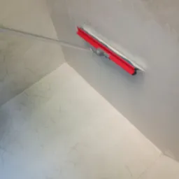 Nakładanie gładzi na ścianę w rogu pomieszczenia za pomocą szpachli z długim uchwytem. Widoczne delikatne wzory na ścianie i podłodze.