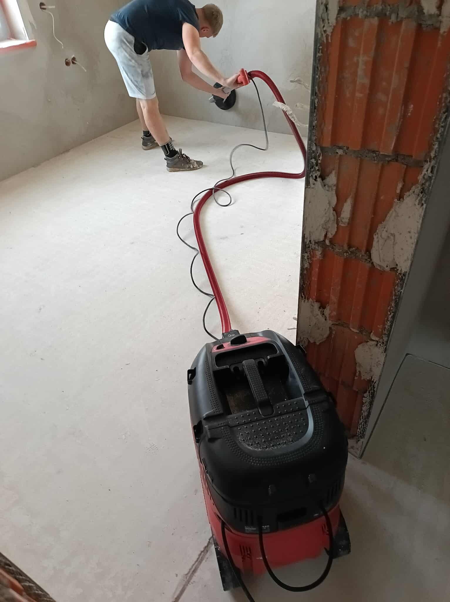 Prace szpachlarskie: mężczyzna szlifuje ścianę szlifierką z odkurzaczem. Widoczna obrobiona ściana i odkurzacz przemysłowy. Ceglany filar w tle.